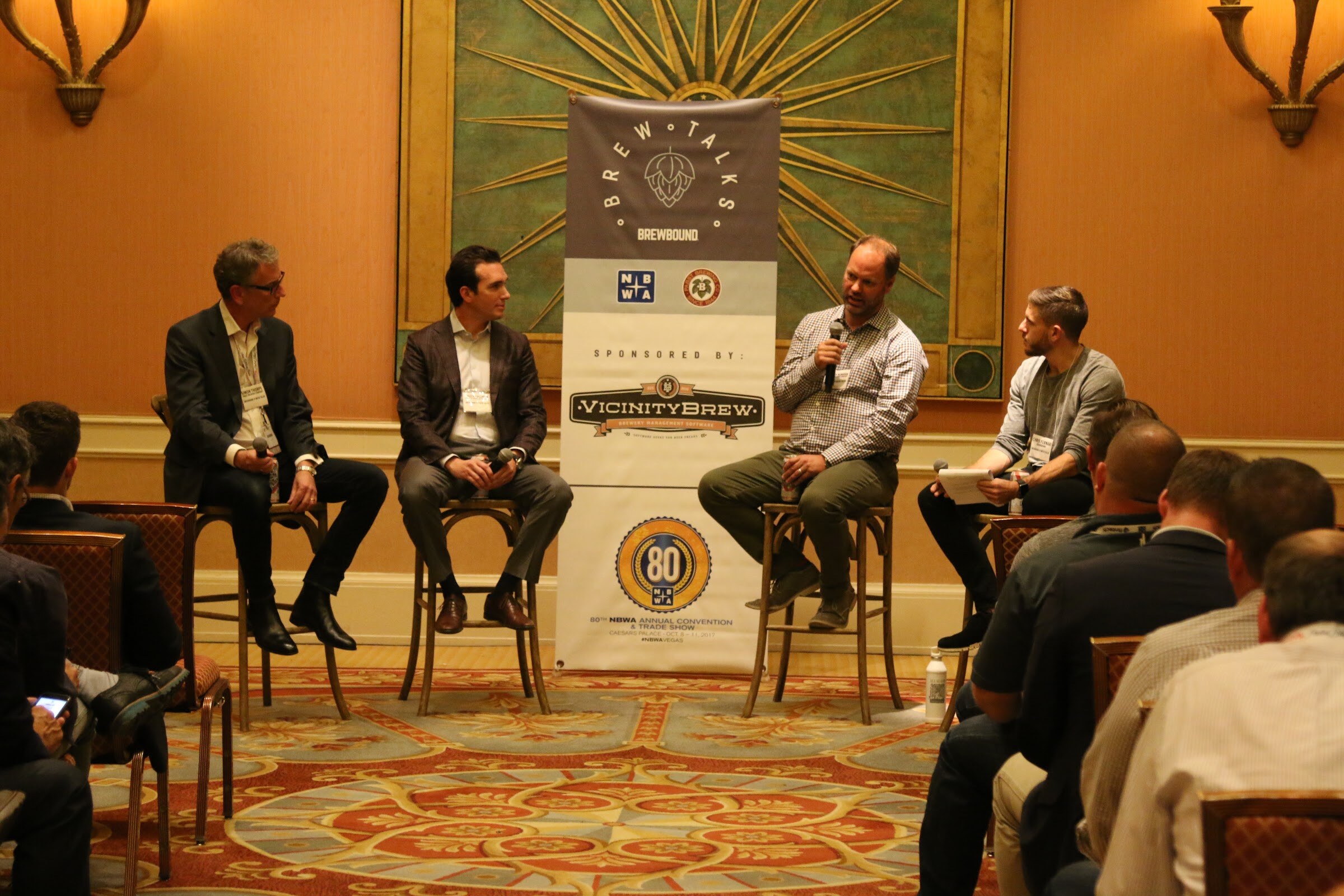 Four men on tall wooden stools in front of a Brew Talks Brewbound banner reading Sponsored by VicinityBrew and 80th NBWA Annual Convention, with seated attendees at the right.