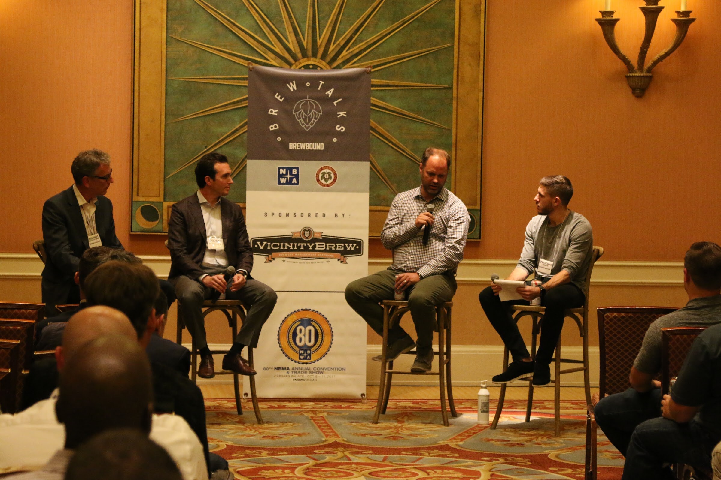 A panel of four seated in front of a Brew Talks banner with NBWA and VicinityBrew logos, with rows of attendees in the foreground.