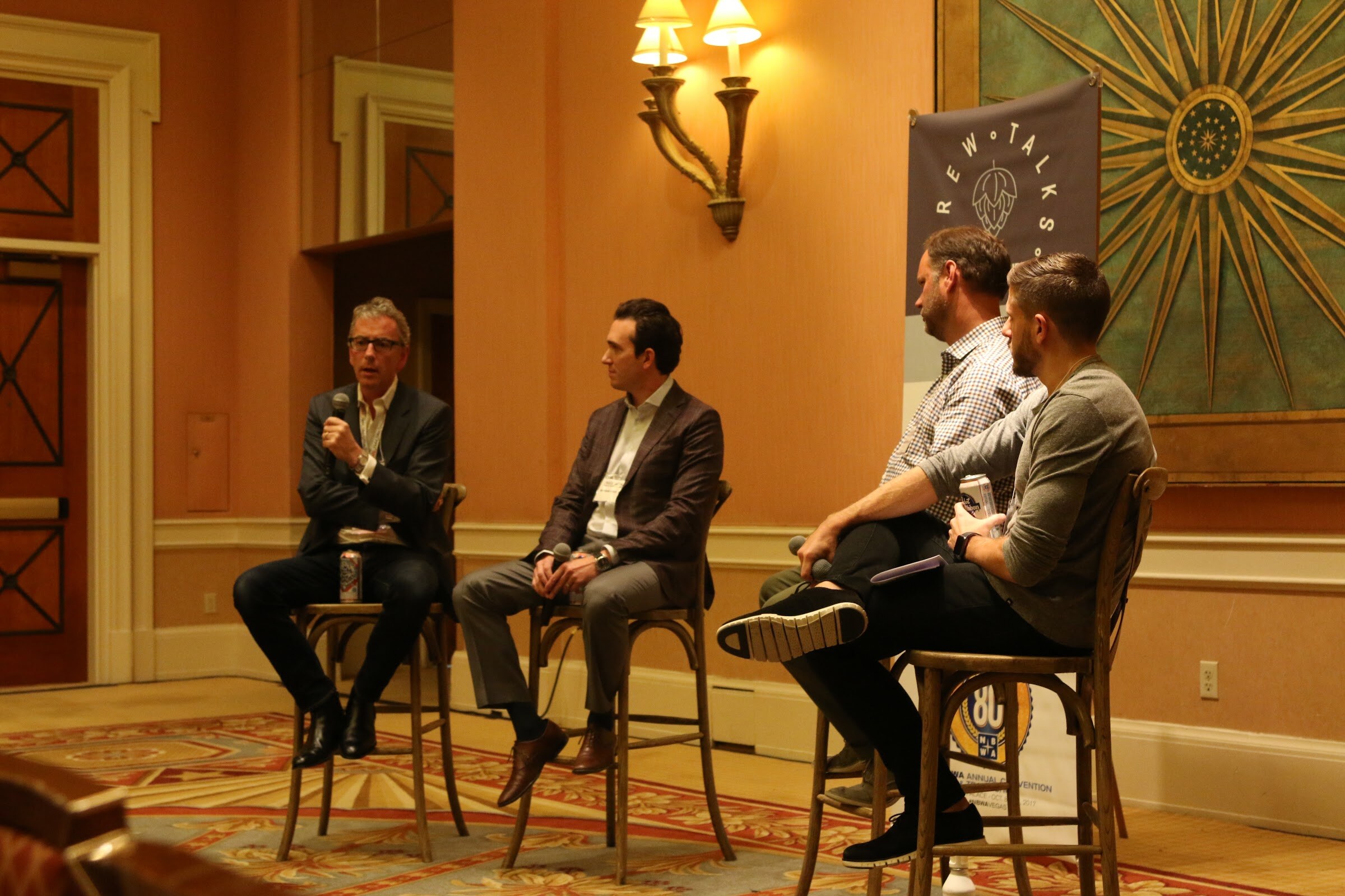 Four men seated on stools on a patterned carpet beside a Brew Talks banner, with the nearest two turned toward each other.
