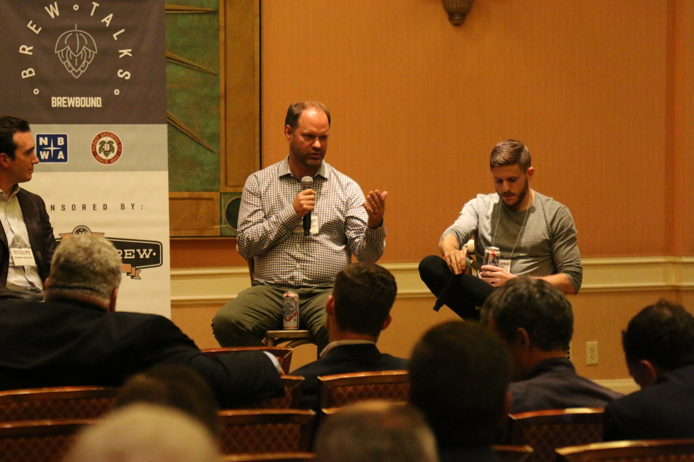 Two men on stools in front of a Brew Talks Brewbound banner, one in a checked shirt speaking into a microphone, with attendees' heads in the foreground.