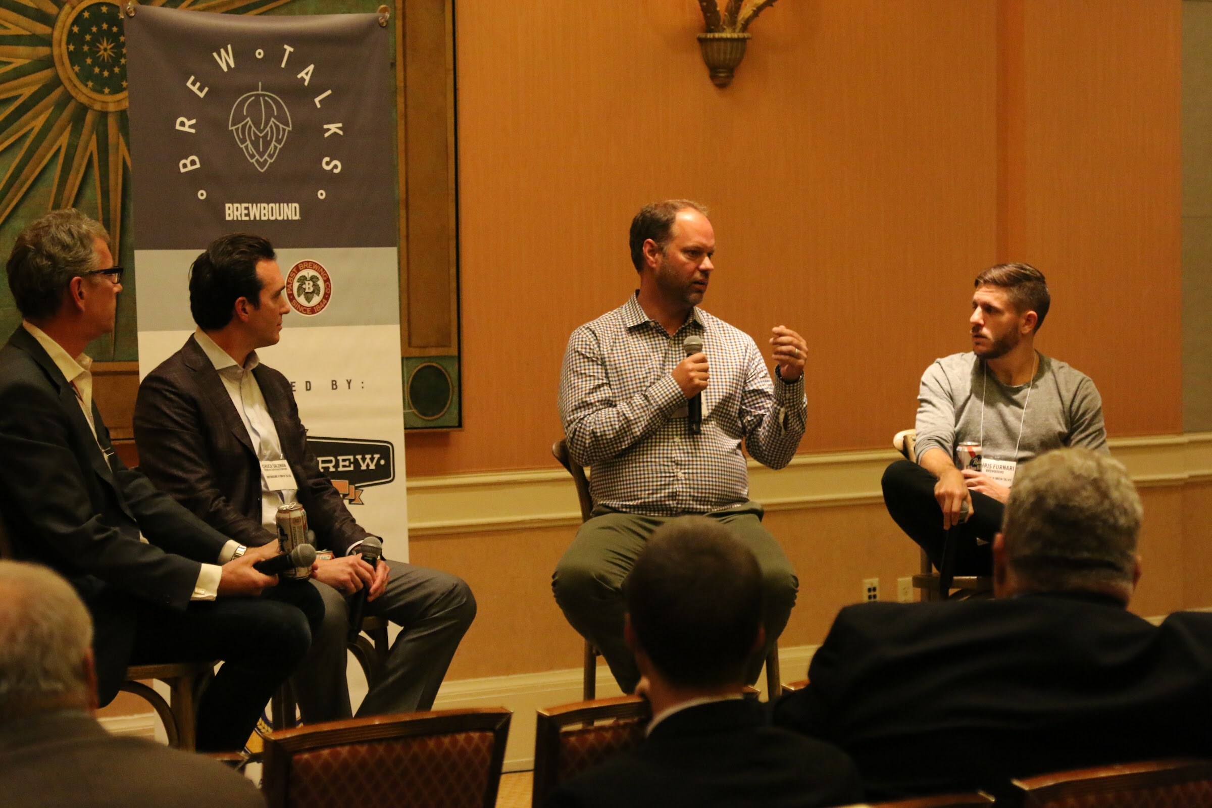 A man in a checked shirt speaks into a microphone as a younger moderator listens, with two men in blazers on stools at the left edge.