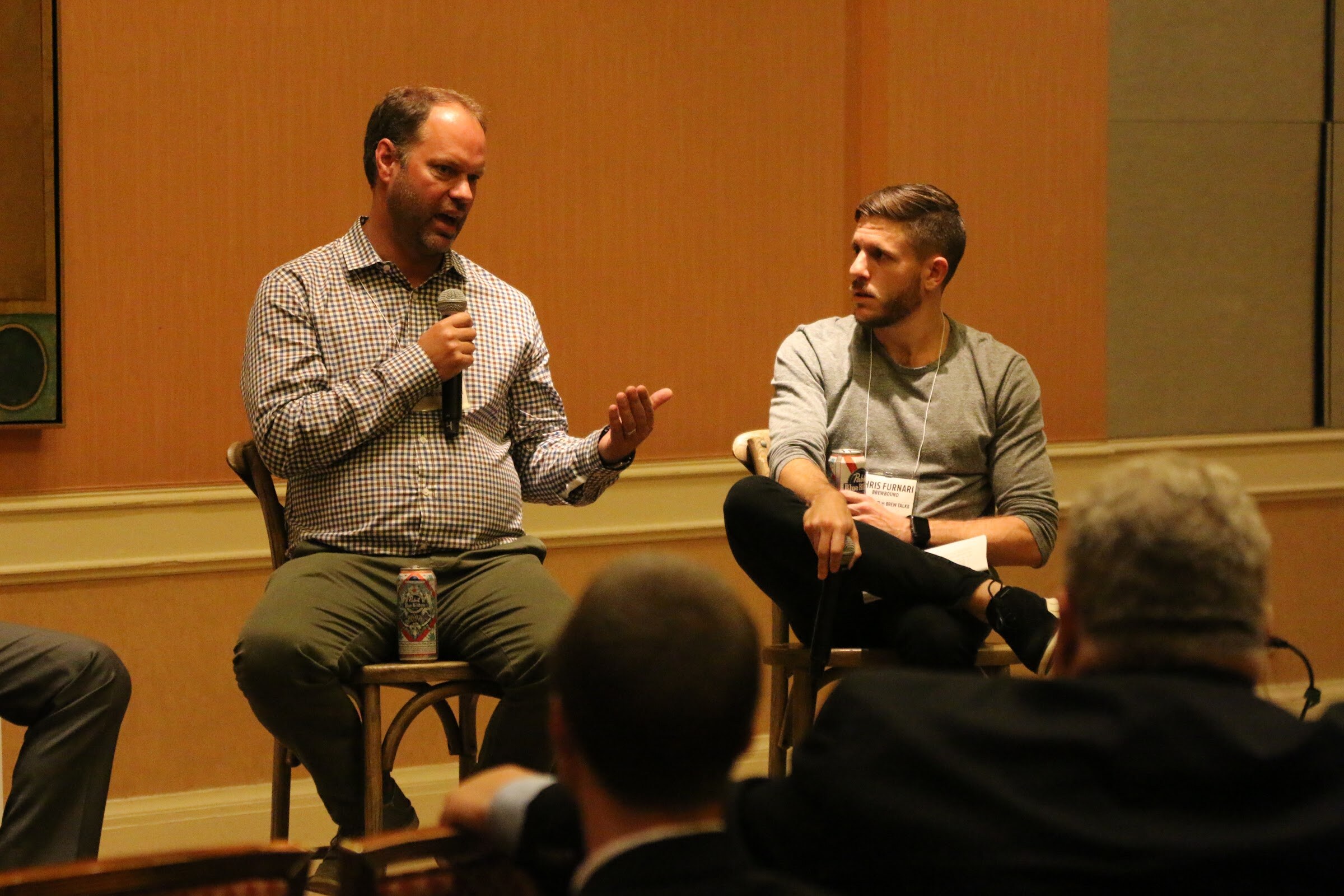 A man in a checked shirt speaks into a microphone while a moderator in gray with a notes sheet watches him, with attendee heads in front.