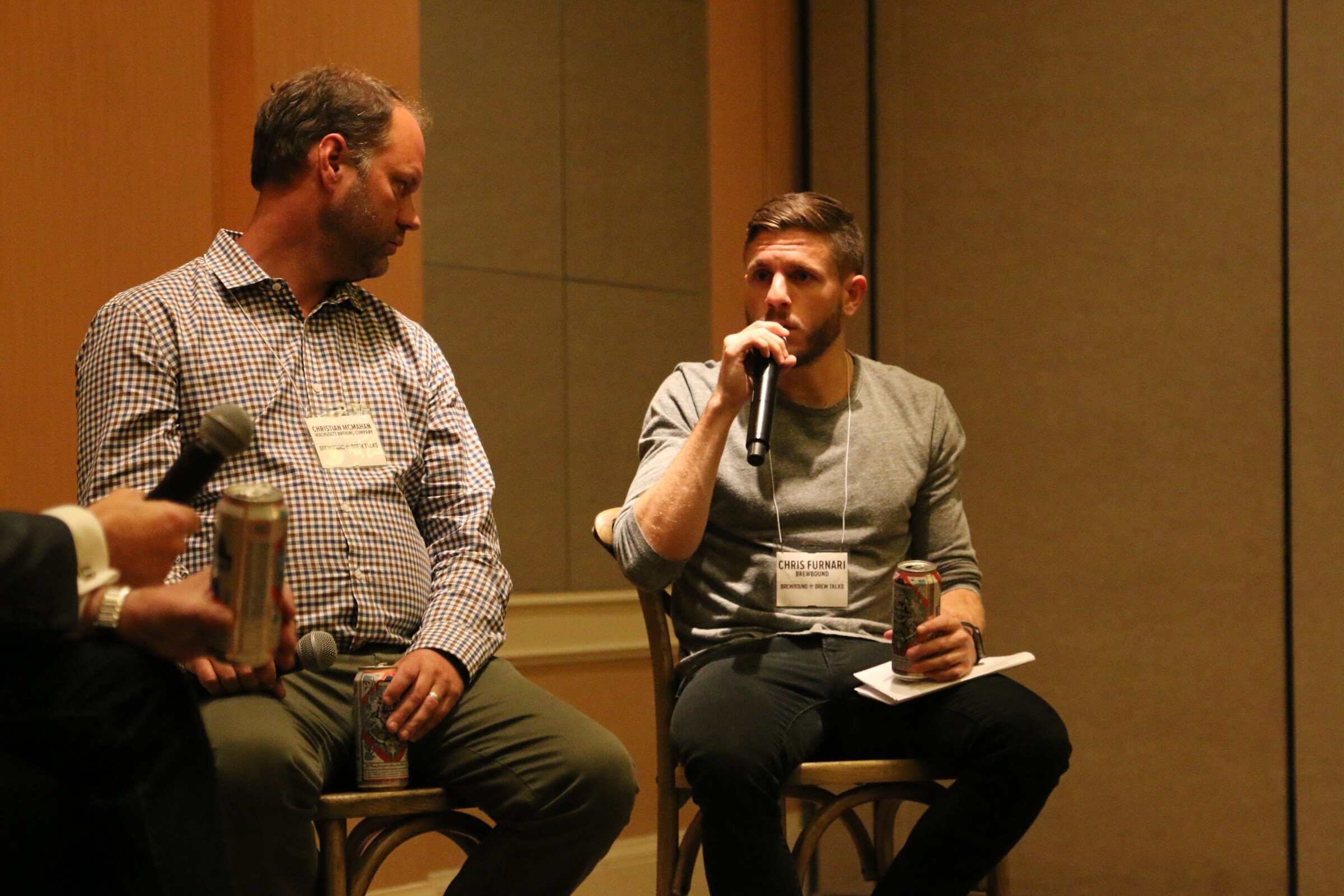 A man in a gray shirt with a conference lanyard speaks into a microphone, with a man in a checked shirt looking down, and a hand holding a can at the left edge.