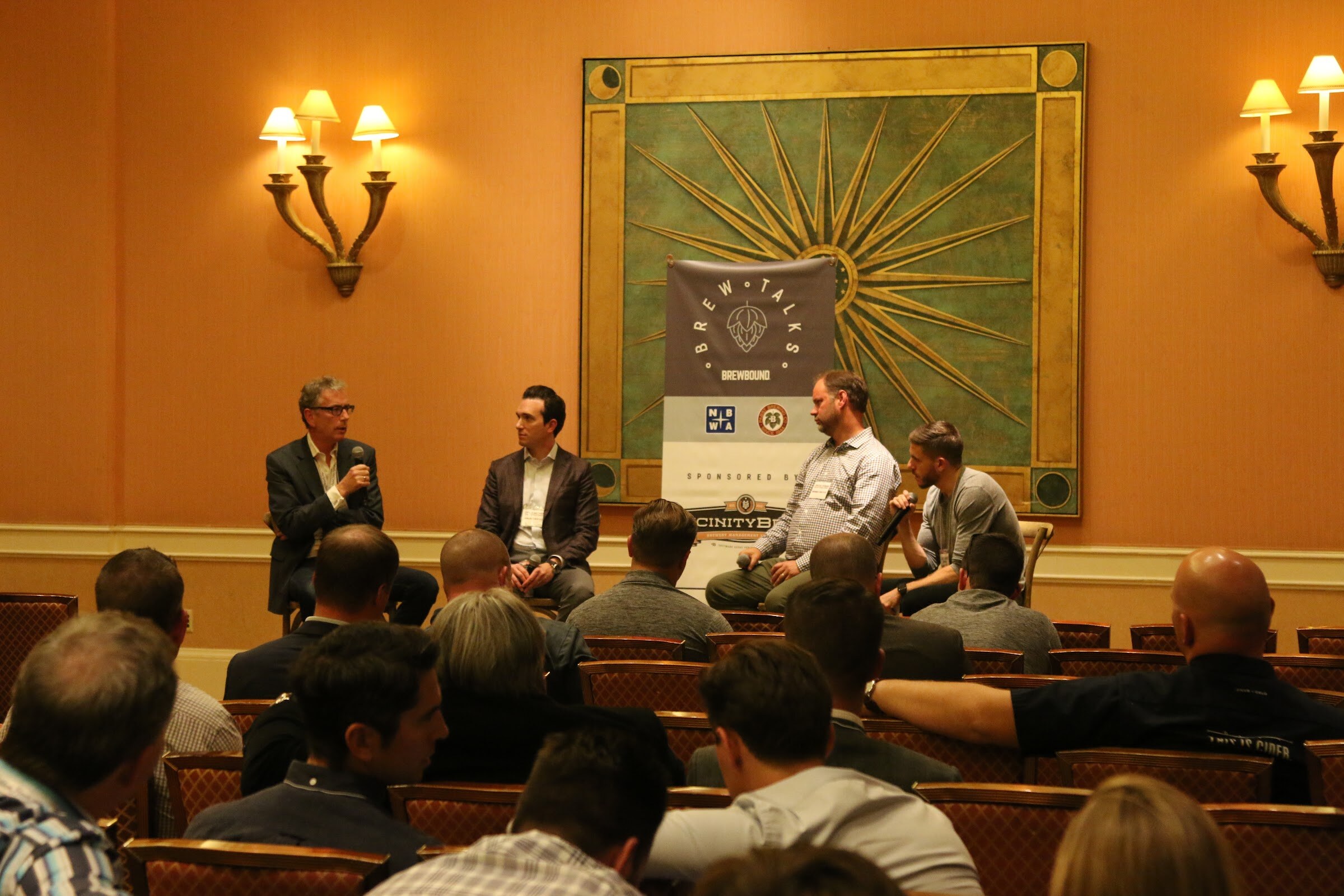 Four panelists on stools beside a Brew Talks banner and a green sunburst wall panel, with attendees in rows facing them.