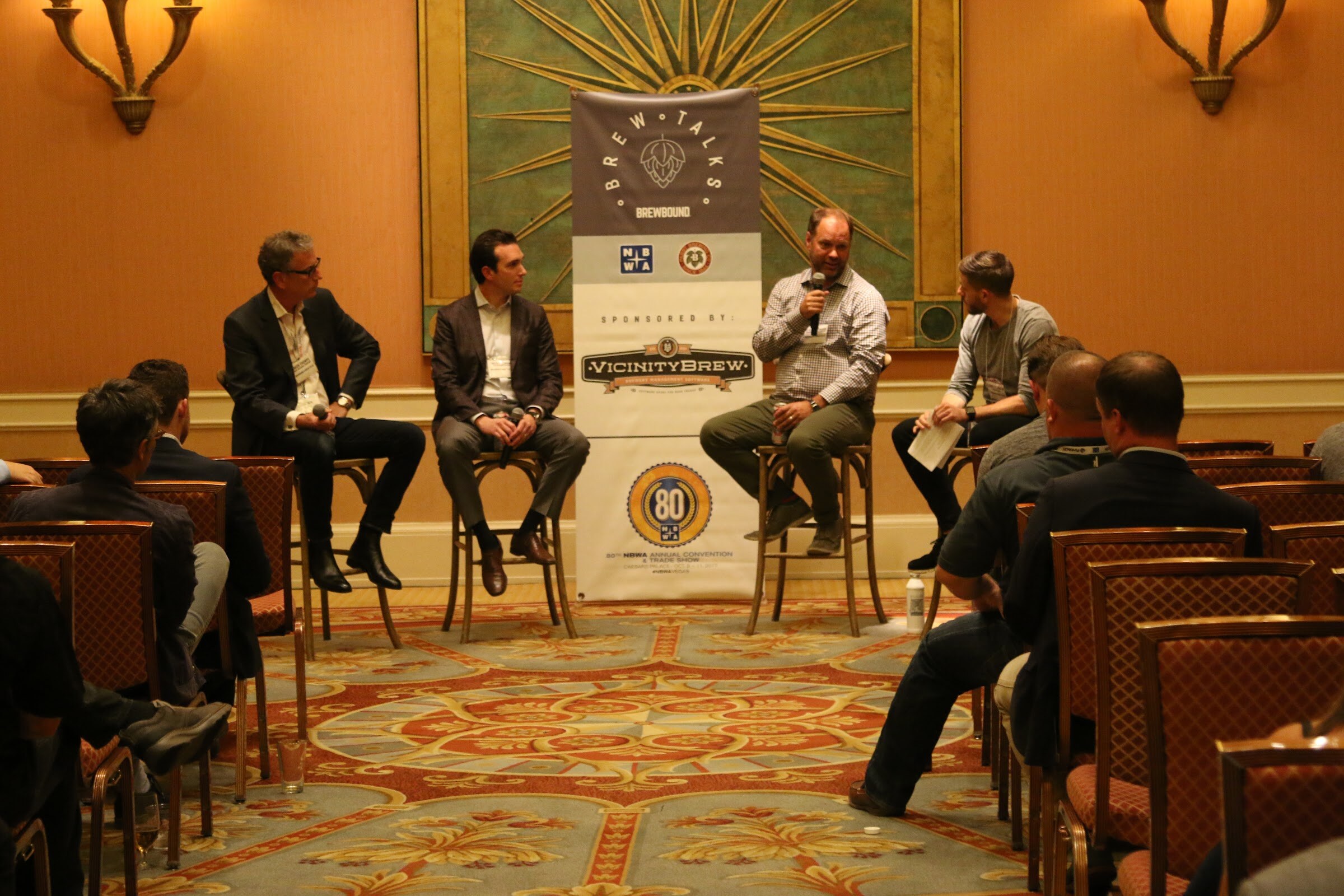 Four men on stools beside a Brew Talks banner and 80th NBWA emblem, with rows of attendees from behind in the foreground.