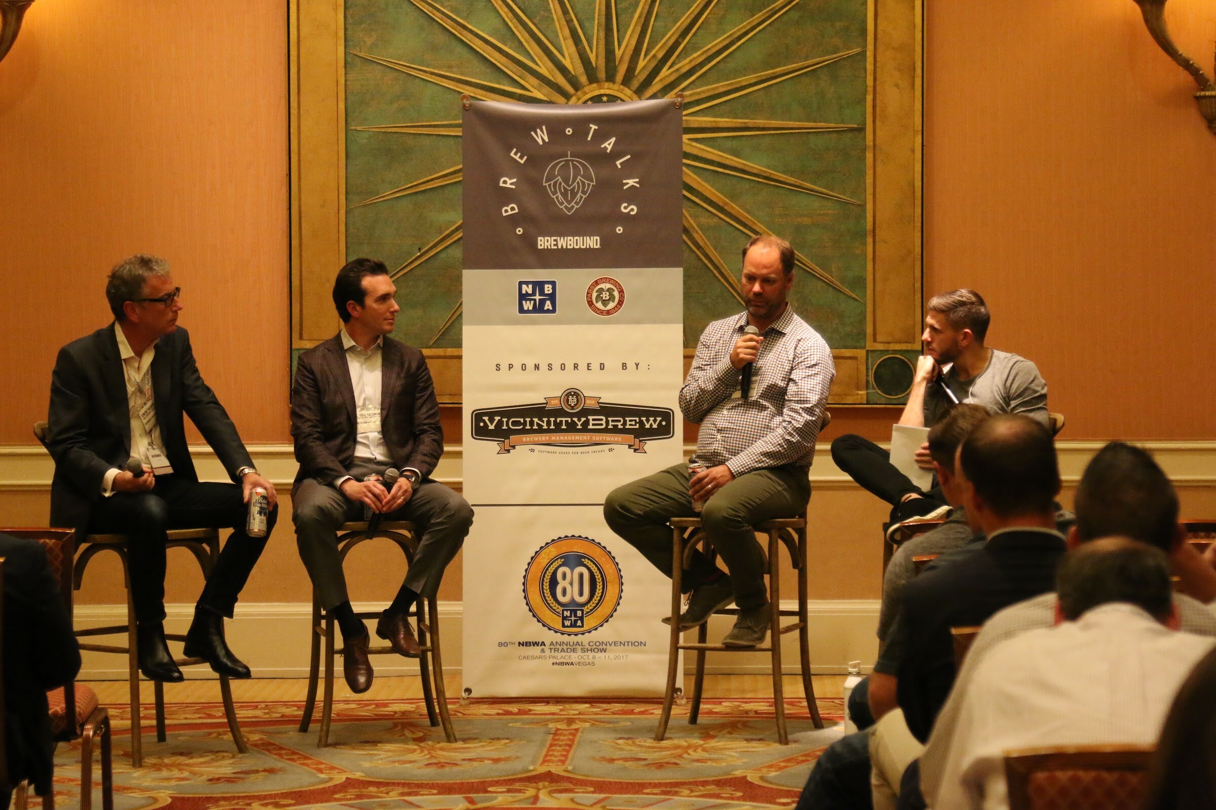 Four seated men on stools at a banner reading Brew Talks and Sponsored by VicinityBrew, with attendees on the right edge.