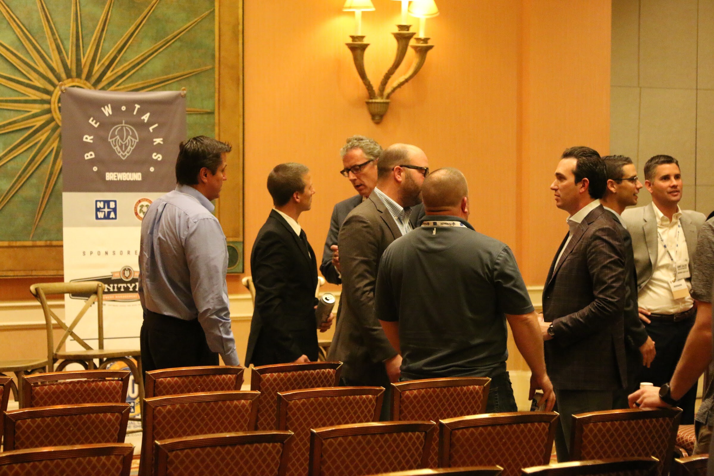 Men in suits and open-collar shirts talking in a group in front of rows of empty chairs and a Brew Talks banner.