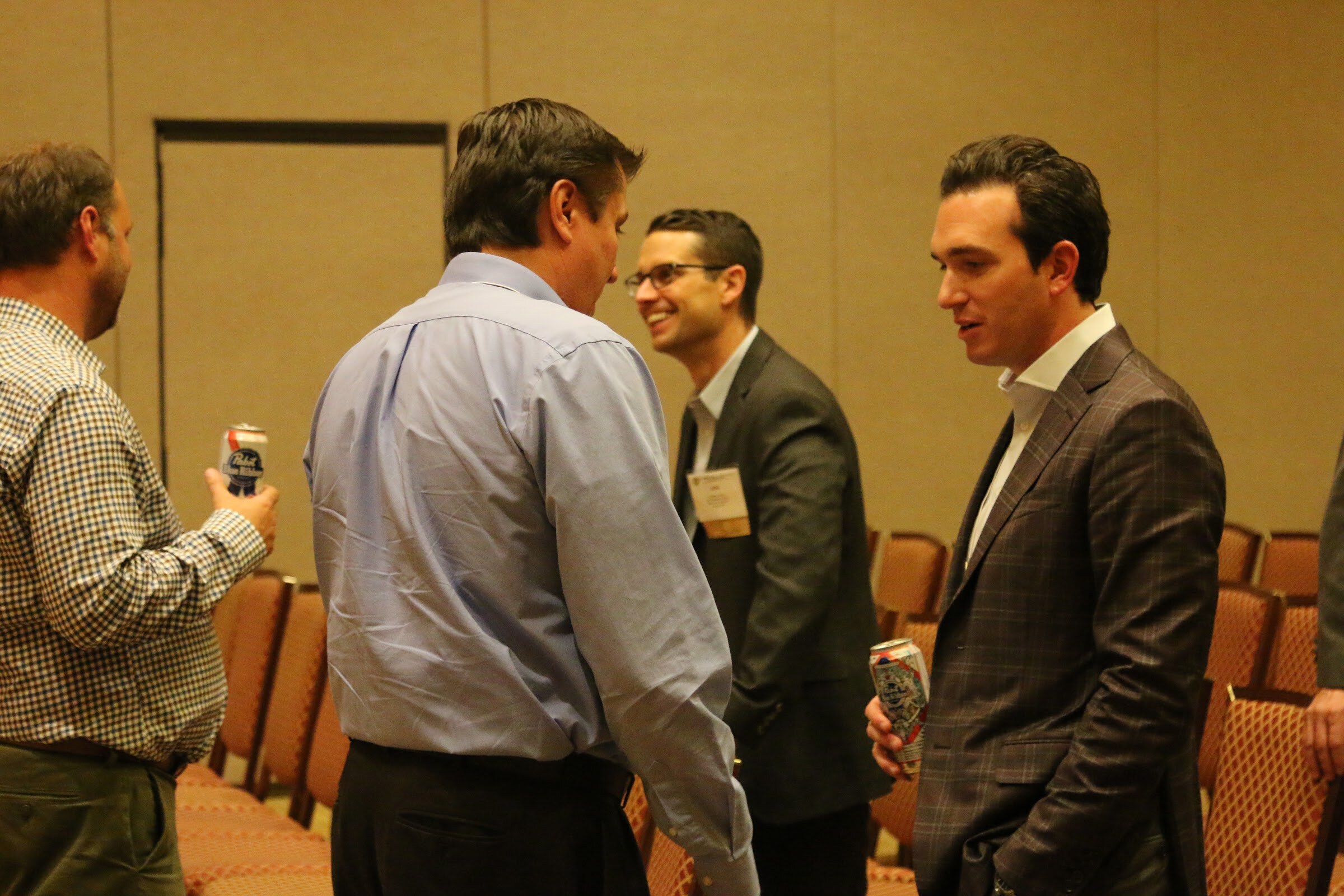 Three men in a meeting room, one holding a can, with a man in a plaid blazer talking with a man in a gray jacket.