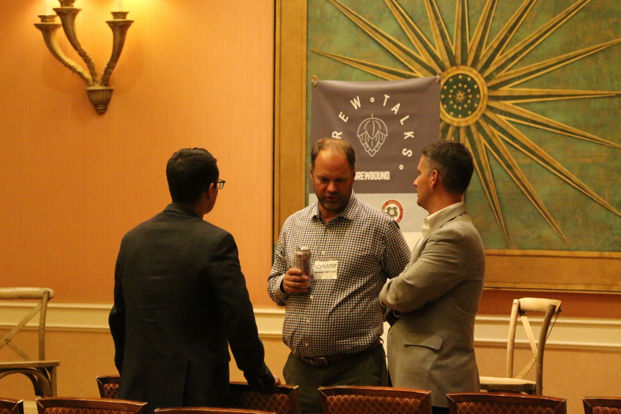 Three men in conversation in front of a Brew Talks banner and a green sunburst panel, one in a checked shirt holding a can.