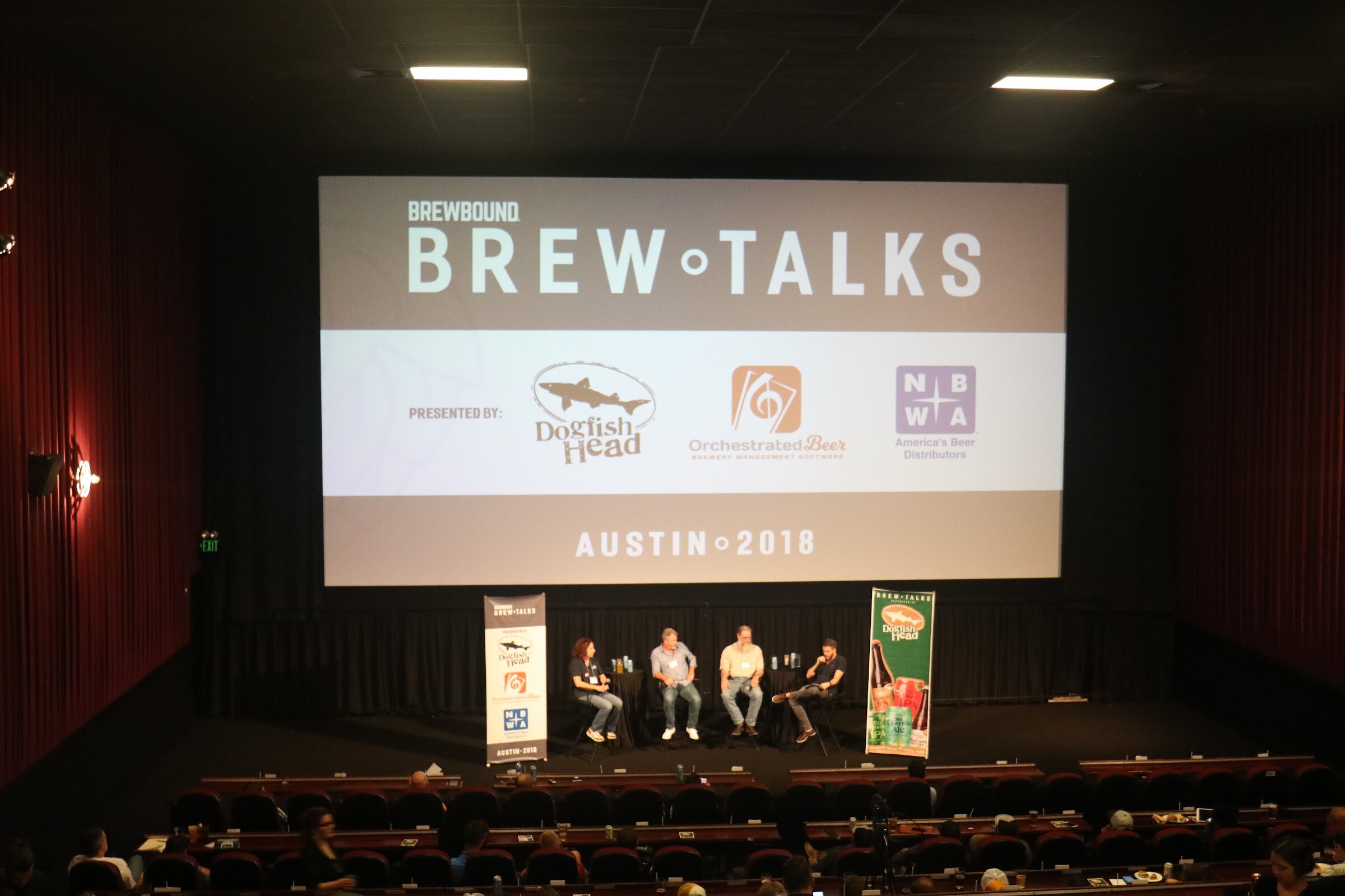 Wide view from the balcony of a movie theater with a large screen reading Brewbound Brew Talks, Presented by Dogfish Head, Orchestrated Beer and NBWA, and four people seated on the stage below.