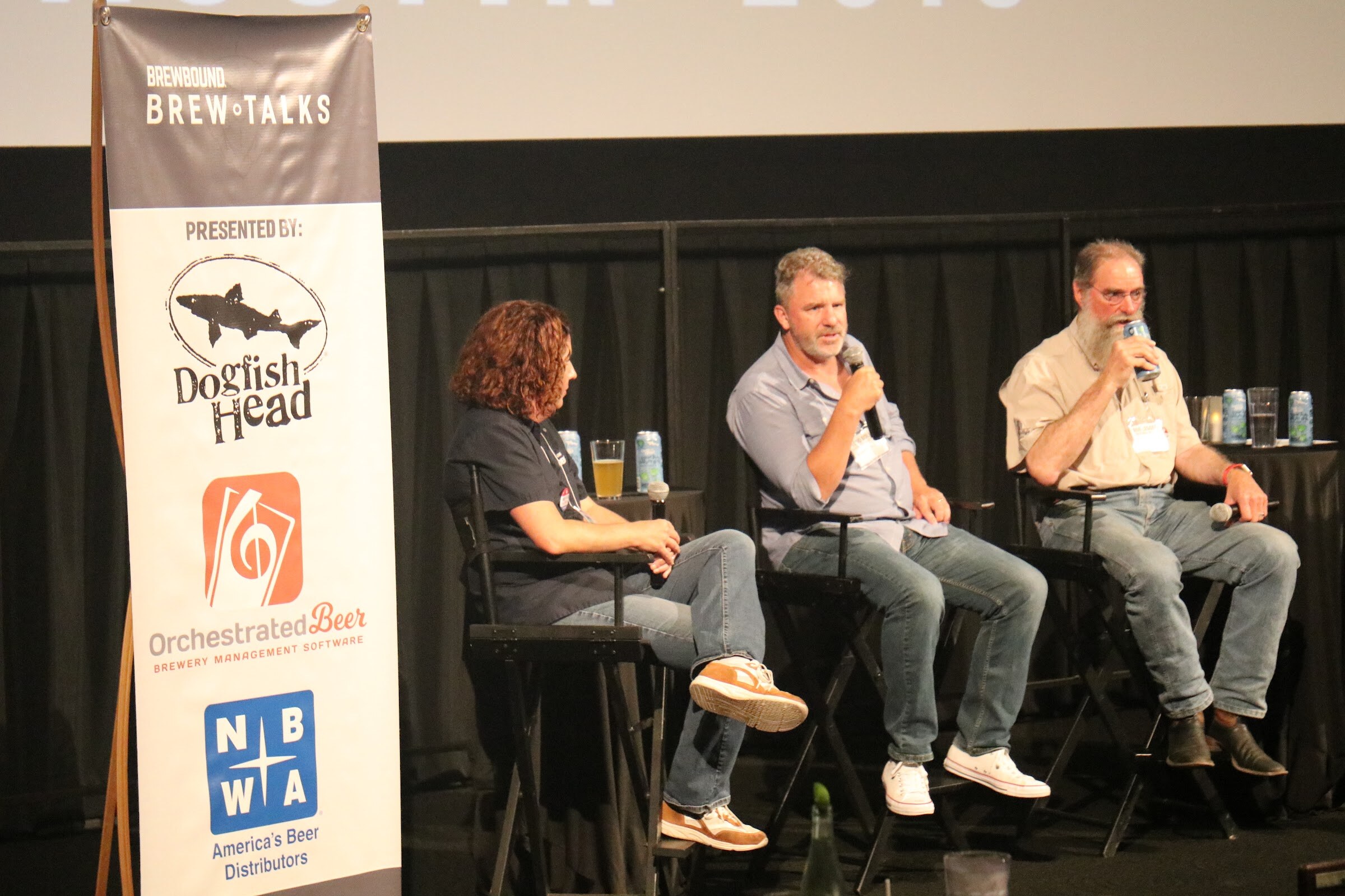 A woman and two men on director chairs on a stage next to a tall banner with Dogfish Head, Orchestrated Beer and NBWA America's Beer Distributors logos.