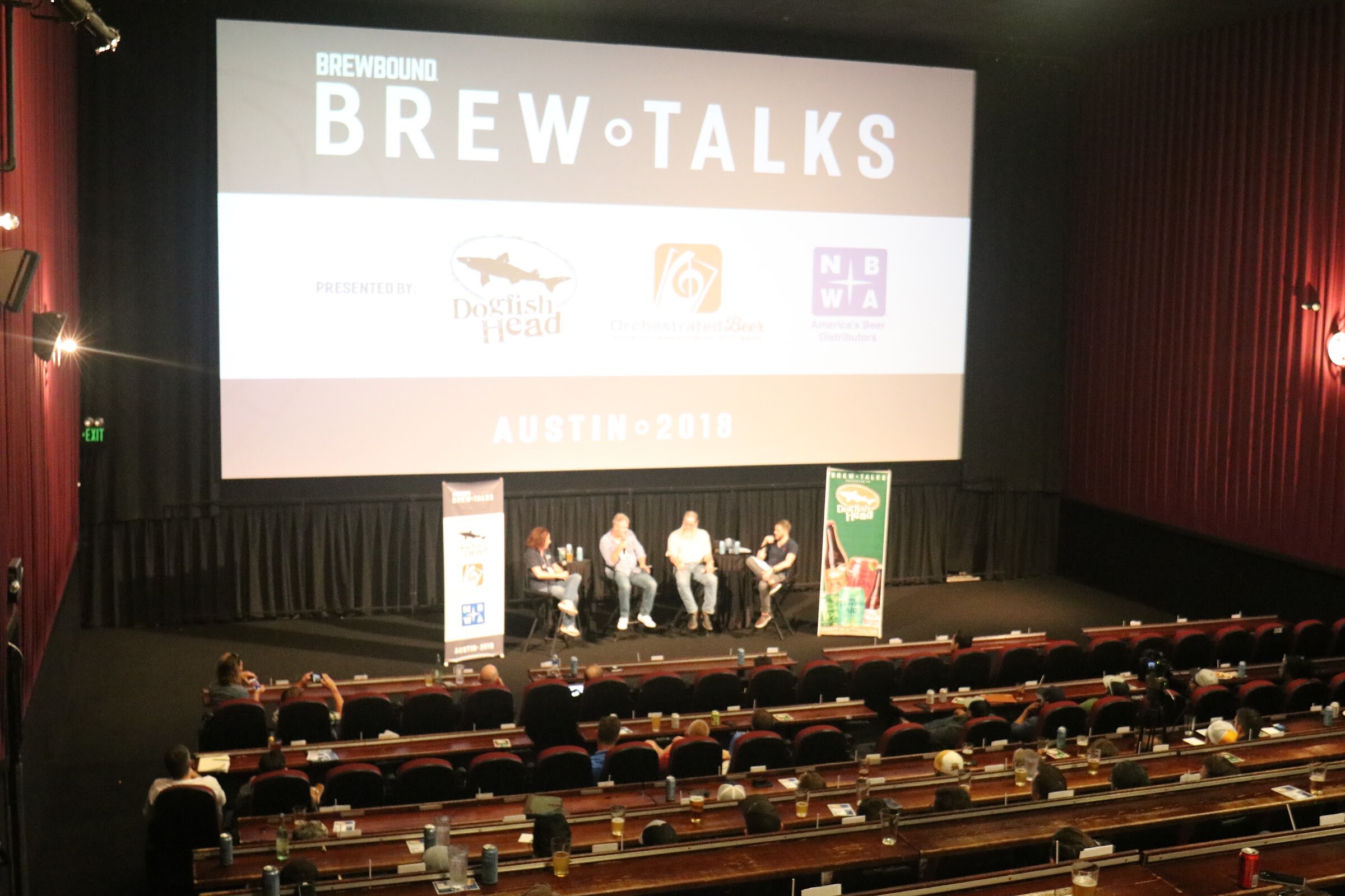 A wide angle of a cinema with rows of red seats and tables, a large Brew Talks screen at the front, and a four-person panel on the stage.