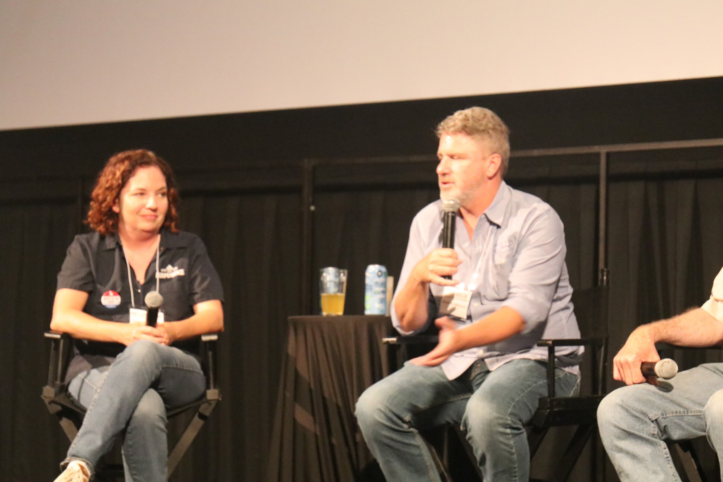 A man in a light blue shirt speaks into a microphone while a woman in a dark polo with a name badge sits smiling beside a small table with a pint of beer and a can.