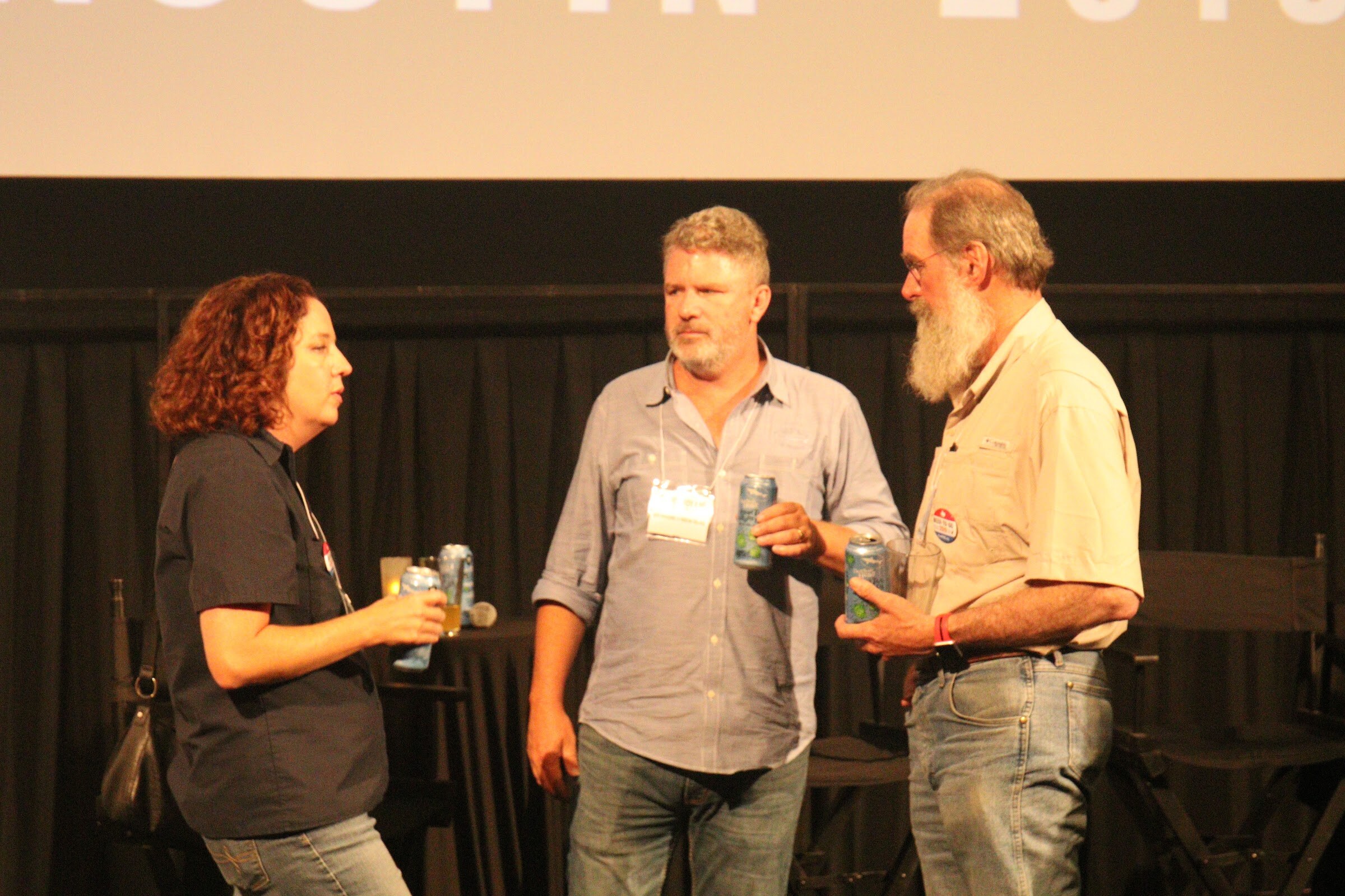 A woman and two men, one with a long white beard, stand talking on a darkened stage holding cans of beer.