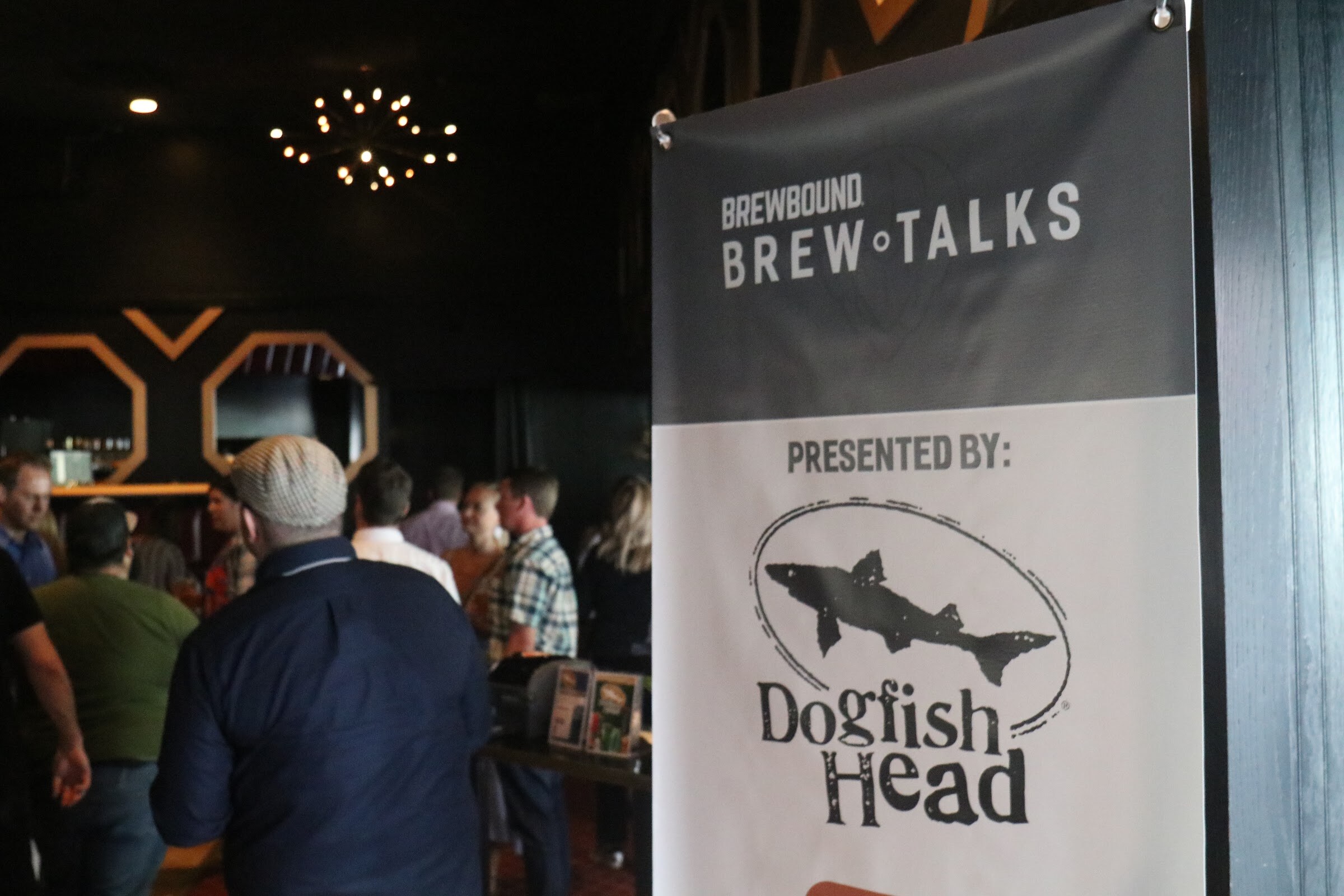 A banner reading Brewbound Brew Talks, Presented By Dogfish Head, with a crowd of guests talking in a dark lobby with a decorative chandelier behind it.