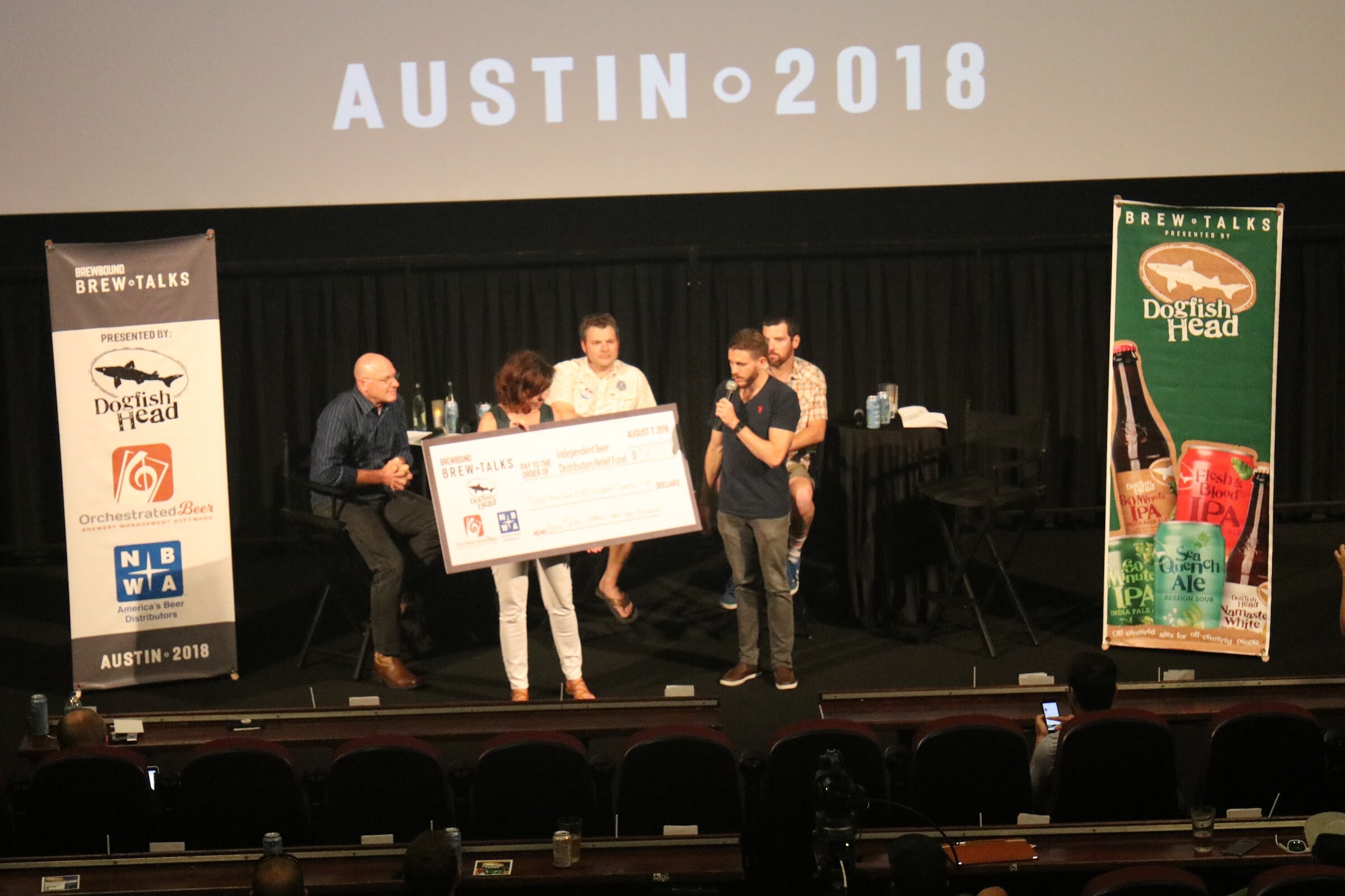 A man with a microphone, a woman and another man hold a big ceremonial check on a stage, with a seated bald man and a bearded man behind them, beneath a screen reading Austin 2018.