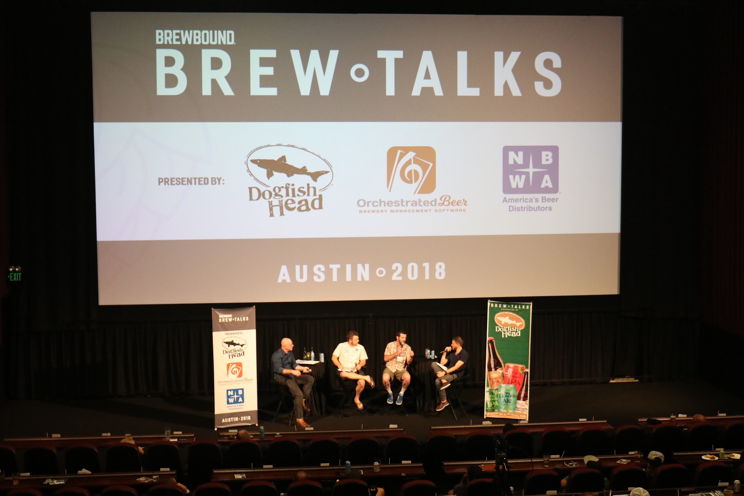 A wide shot from above of a cinema stage where a bald man, two men in casual shirts and a moderator sit on stools below a screen showing the Brew Talks sponsors.