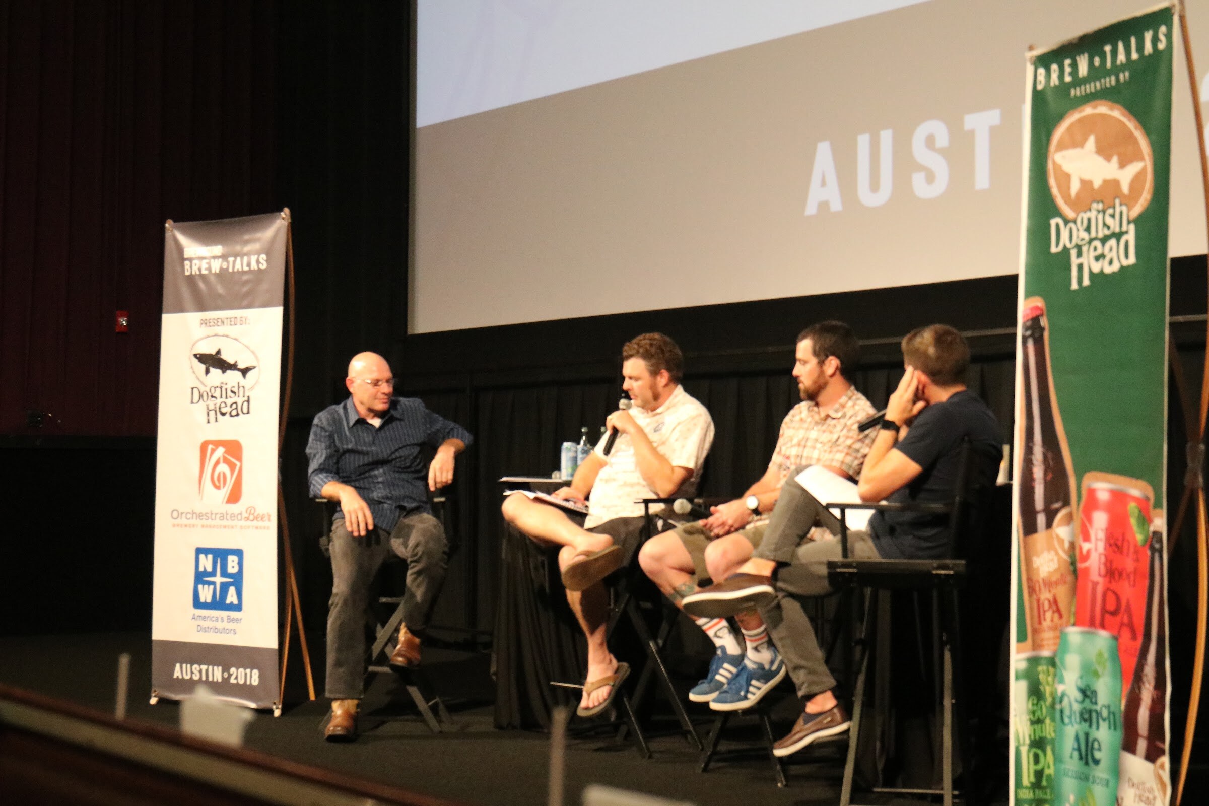 Four men sit on director chairs on a theater stage between a Brew Talks banner with Dogfish Head, Orchestrated Beer and NBWA logos and a green Dogfish Head banner.
