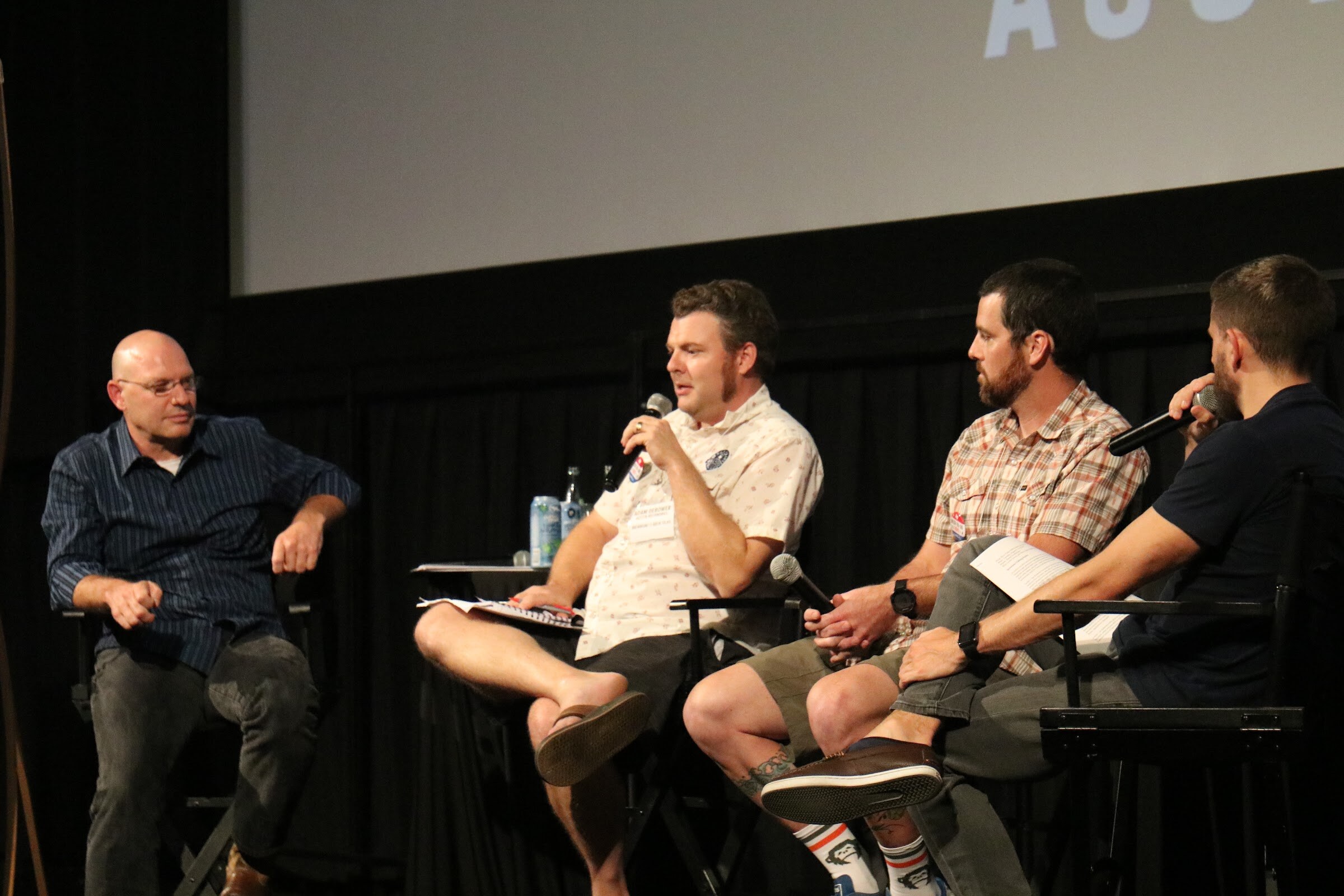 Four men on stage: a bald man in glasses sits back at left, a man in a cream shirt speaks into a microphone, a bearded man in a plaid shirt listens, and a moderator holds a microphone.