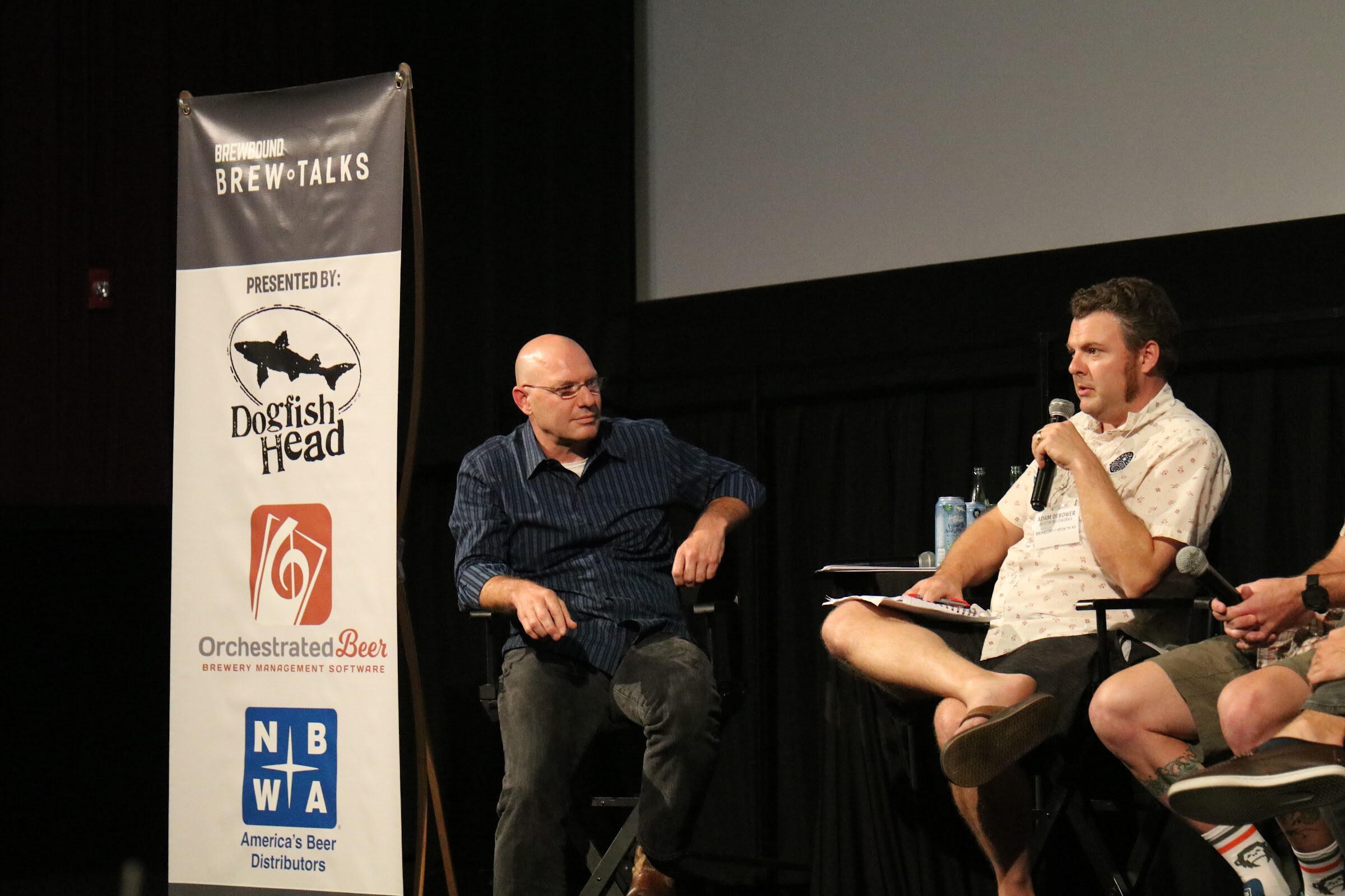 A bald man in a navy striped shirt sits beside a tall banner for Dogfish Head, Orchestrated Beer and NBWA while a man in a patterned shirt speaks into a microphone.