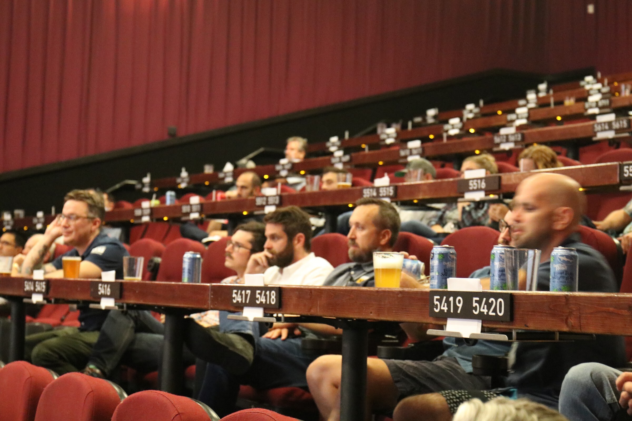 Rows of seated attendees with pints and cans at long wooden tables in a movie theater with red seats and curtains.