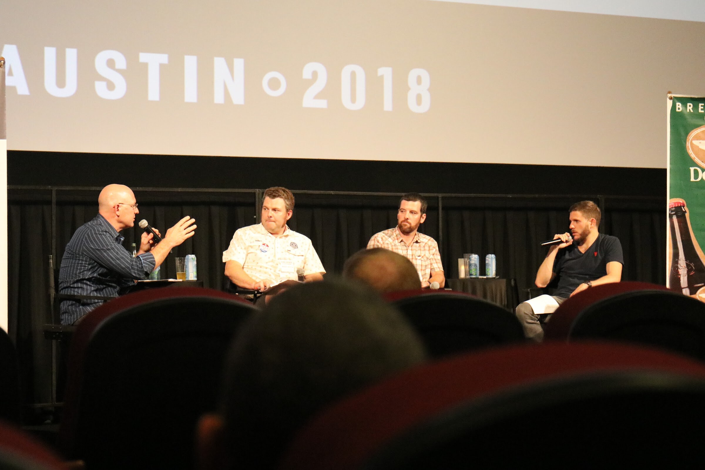 Four men on stage under a screen reading Austin 2018, one gesturing at left with a microphone, with the backs of theater seats in the foreground.