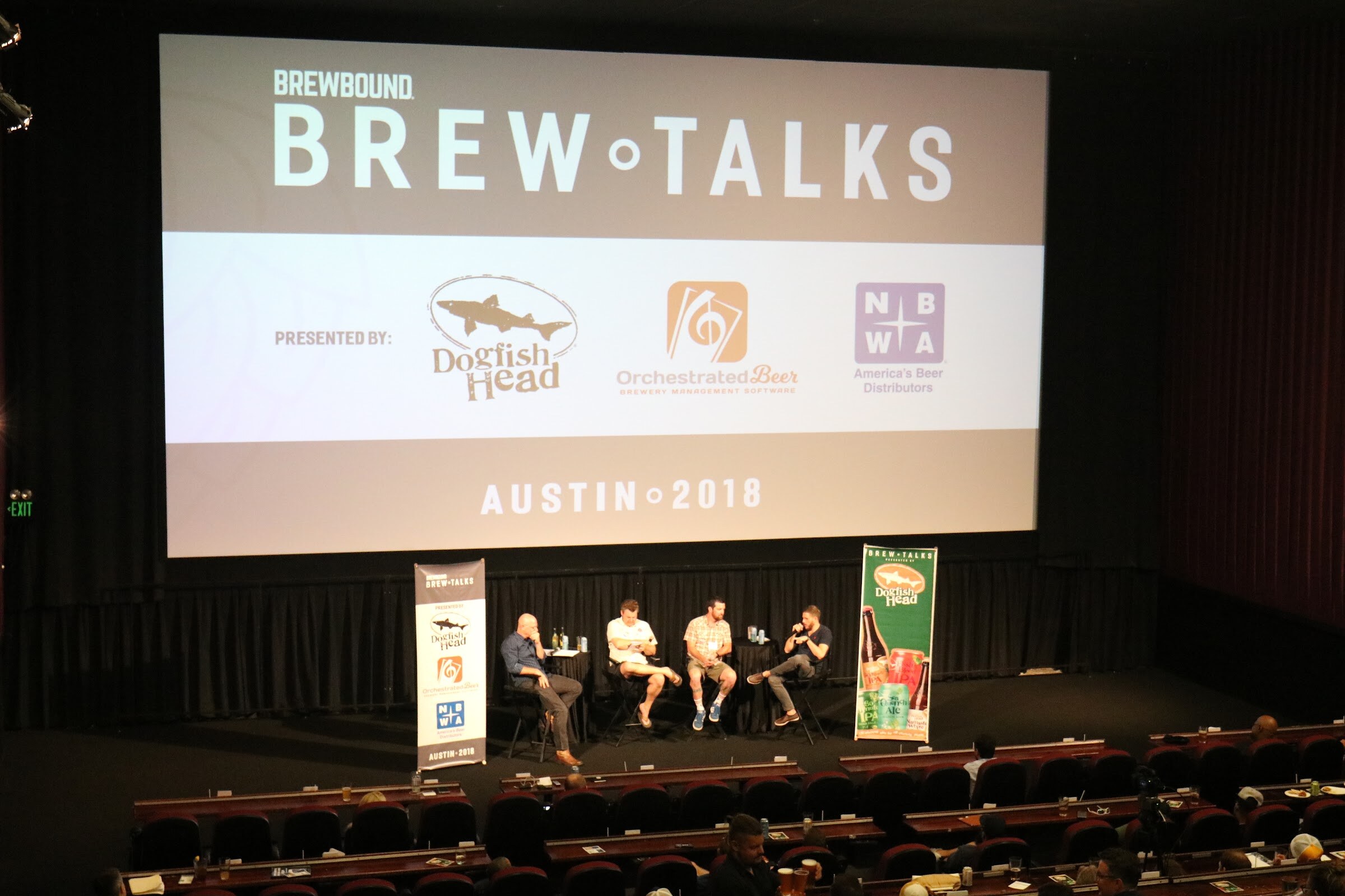 A cinema stage with four people on stools beneath a large Brewbound Brew Talks screen and banners for Dogfish Head and NBWA, with attendees at tables below.