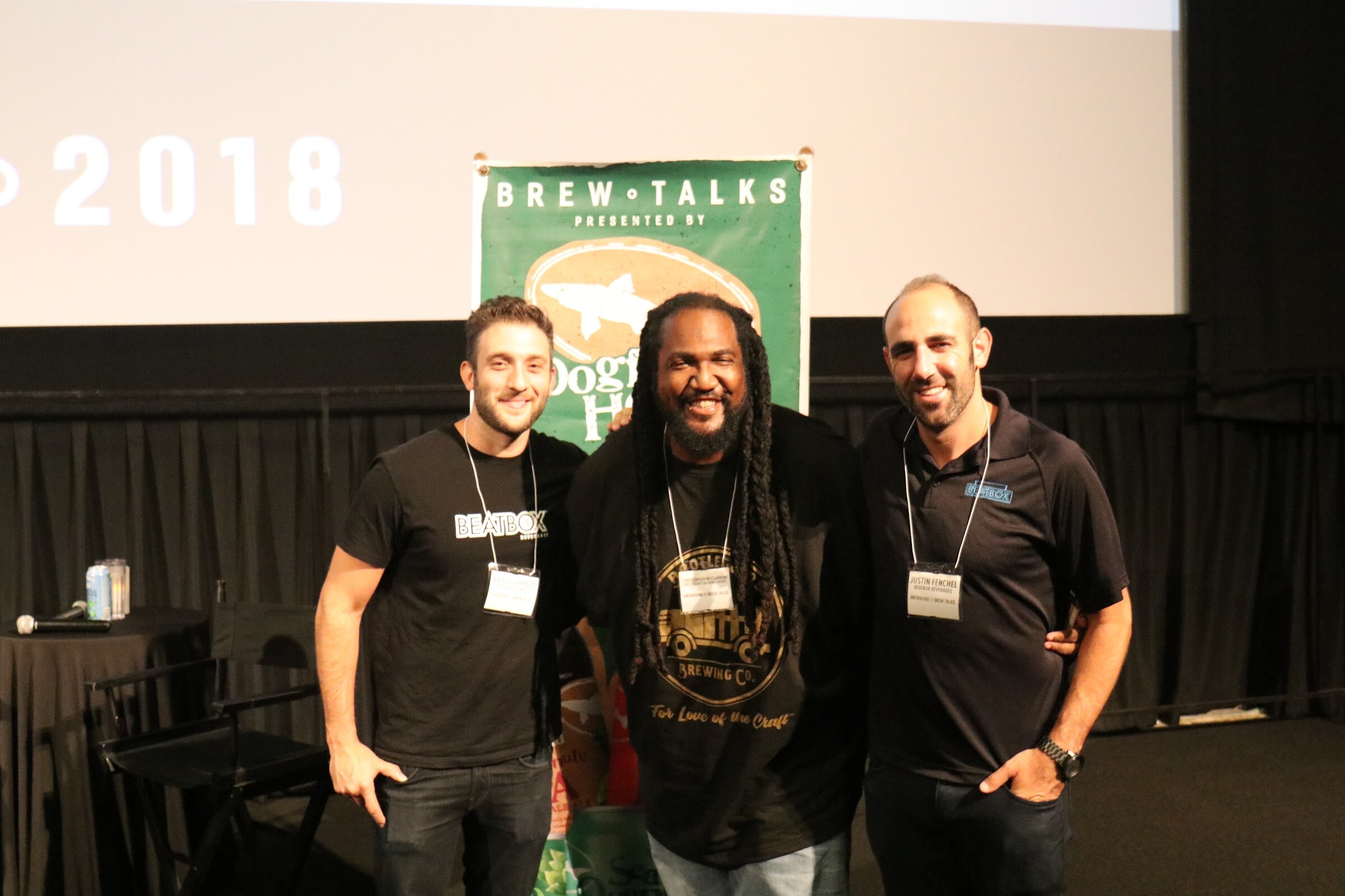 Three men in black shirts with name badges, one with a Beatbox logo and one with a brewing company logo, stand with their arms around each other in front of a green Brew Talks banner.