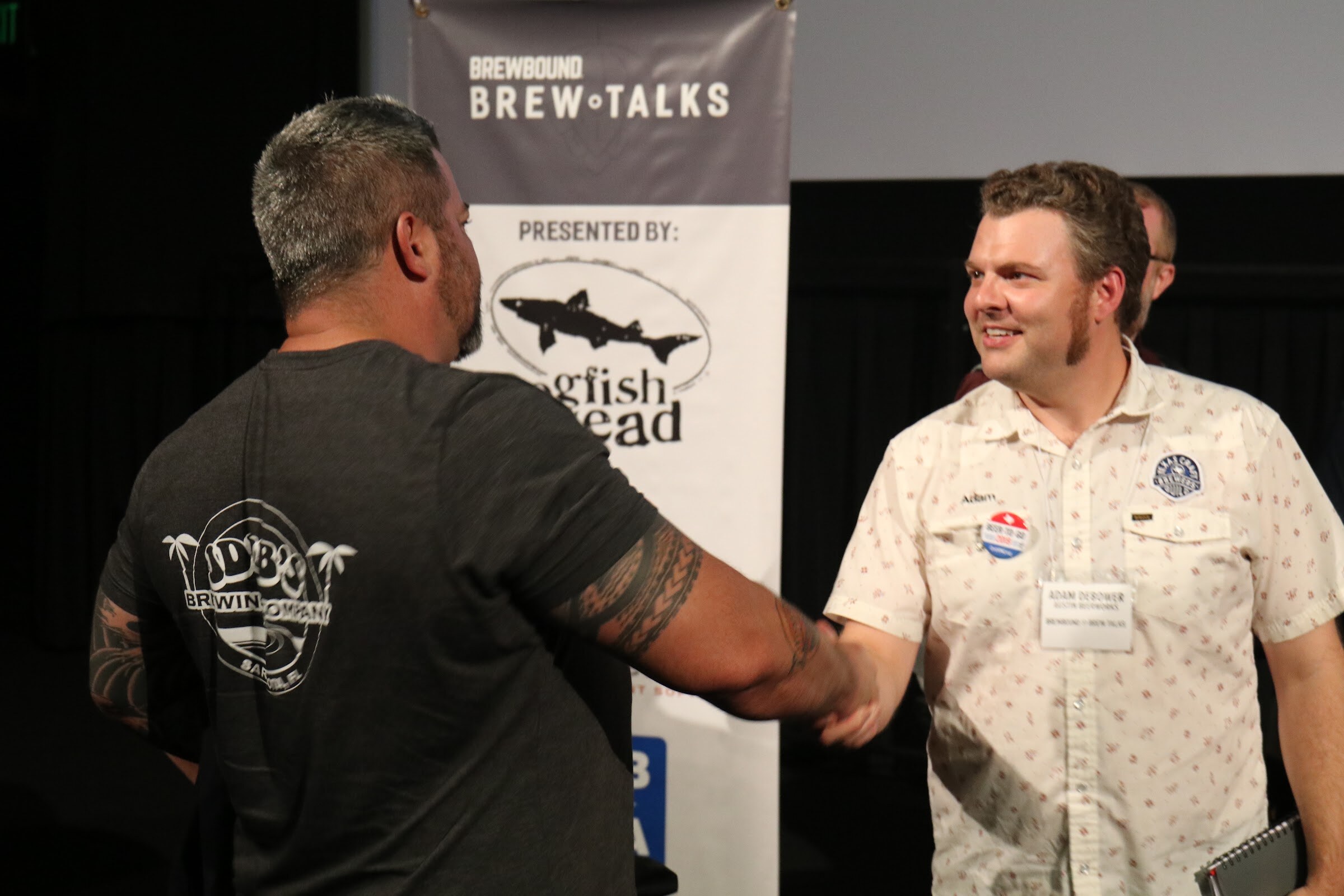 A man in a dark JDub's Brewing Company shirt seen from behind shakes hands with a smiling man in a patterned white shirt and name badge, in front of a Dogfish Head banner.