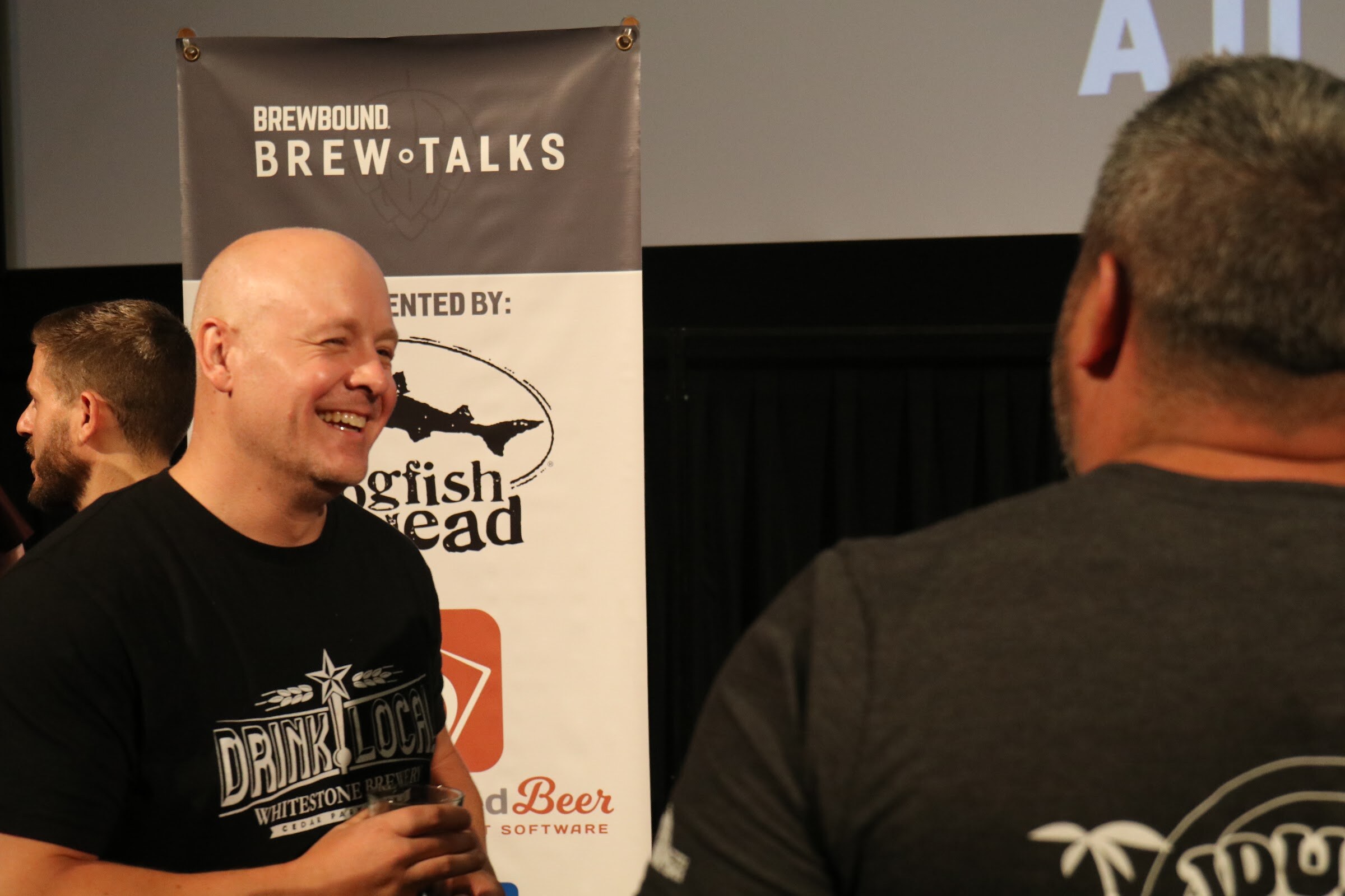 A bald man laughing in a black Whitestone Brewery Drink Local shirt, with another man in a JDub's shirt seen from behind in the foreground and a Dogfish Head banner behind.