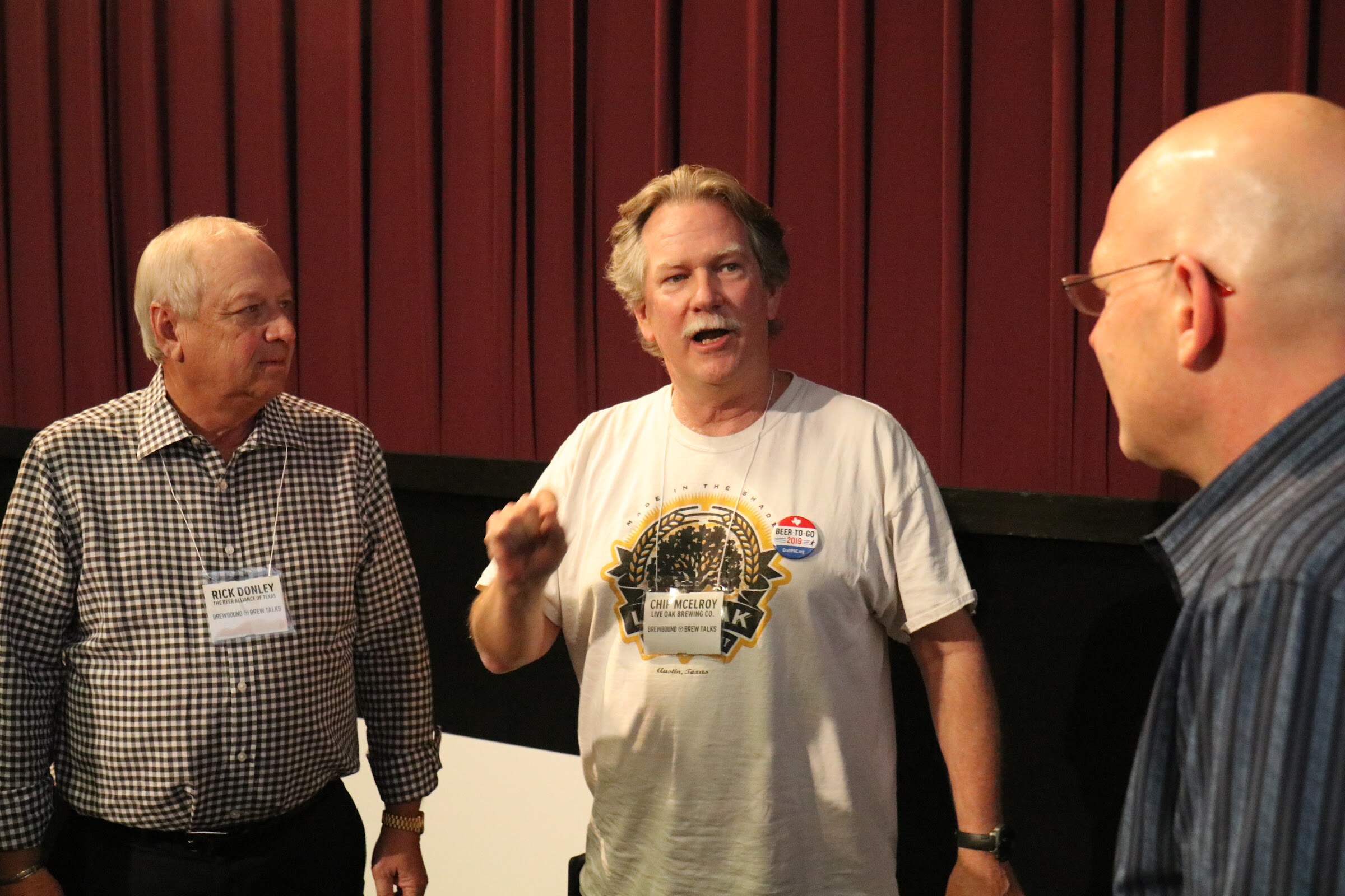 A man in a checked shirt, a man in a white brewery T-shirt gesturing as he speaks, and a bald man in glasses in profile stand together in front of red theater curtains.