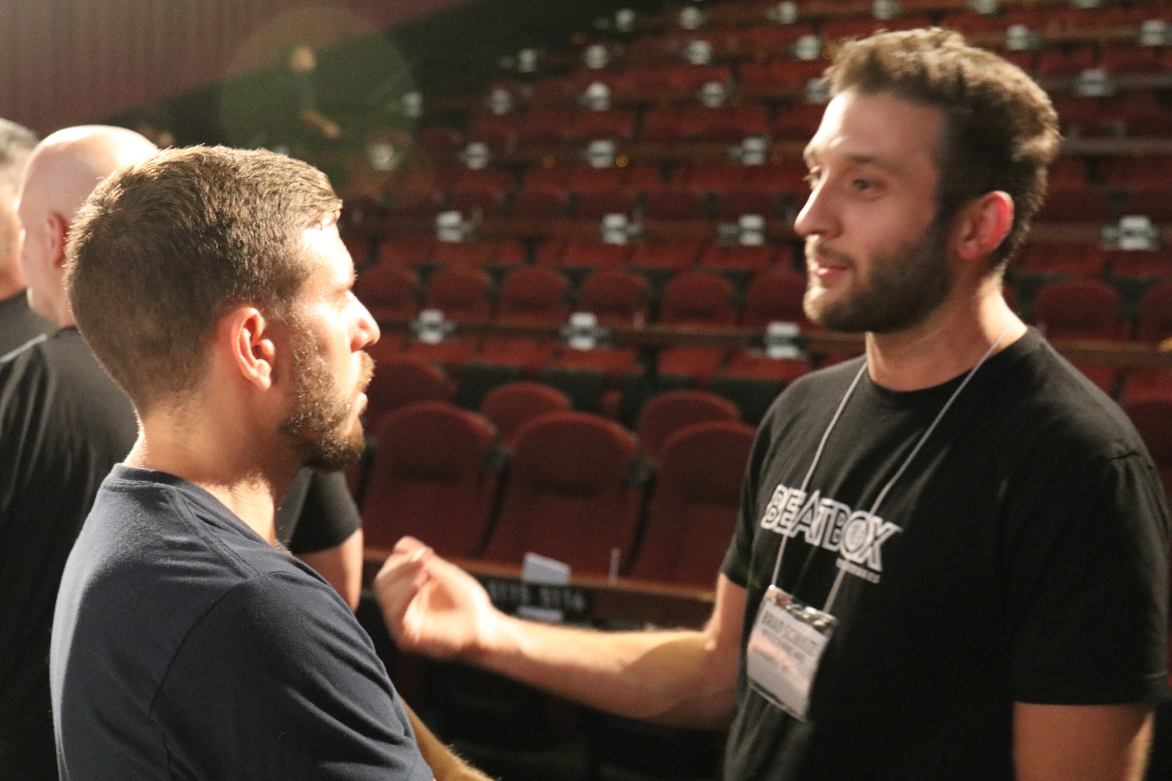 A bearded man in a black Beatbox T-shirt with a name badge talks with a man in a navy shirt seen from the side, with rows of red cinema seats behind them.