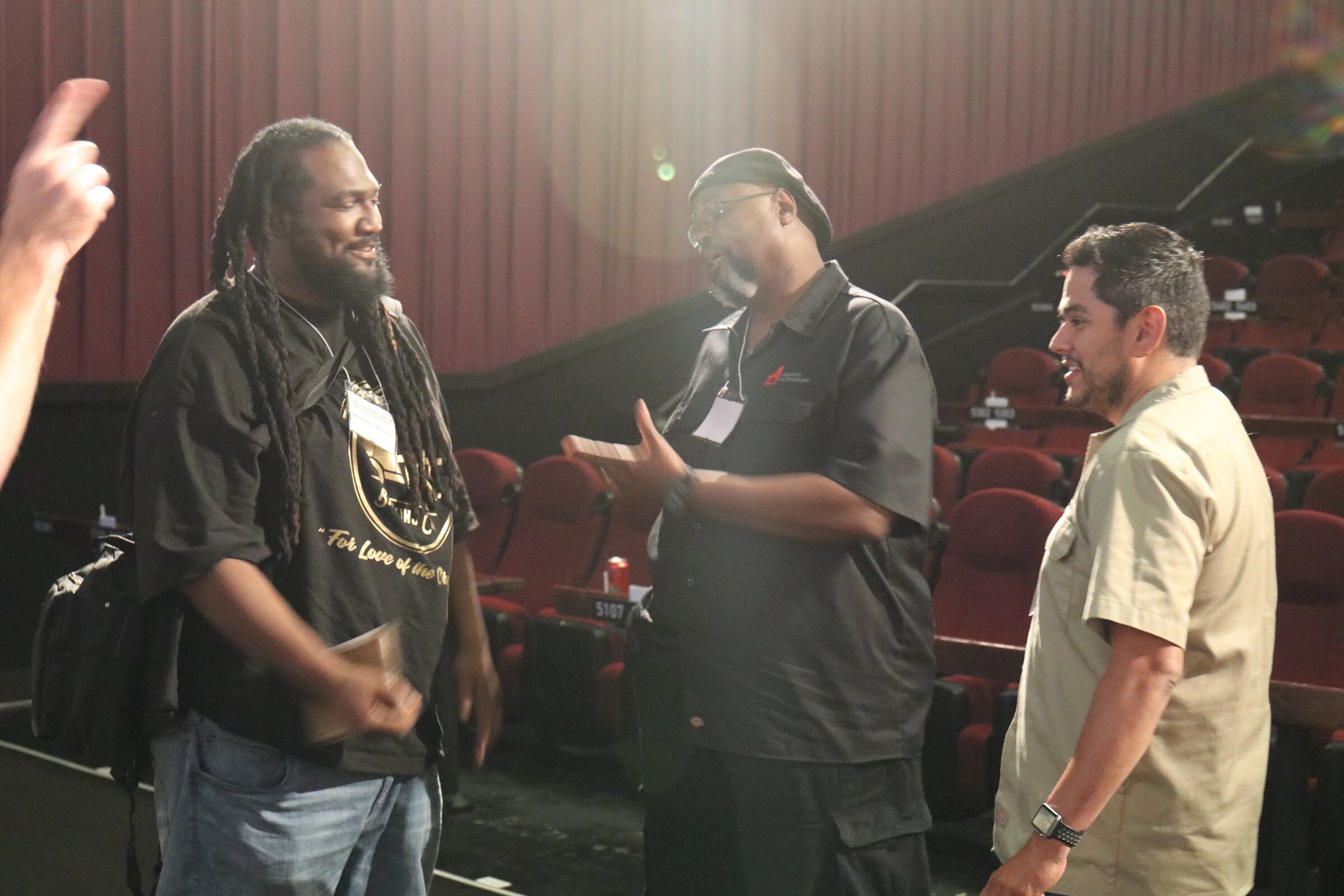 A man in a black brewery T-shirt, a man in a black polo with a cap and a man in a tan shirt talk in a theater with red curtains and rows of seats behind them.