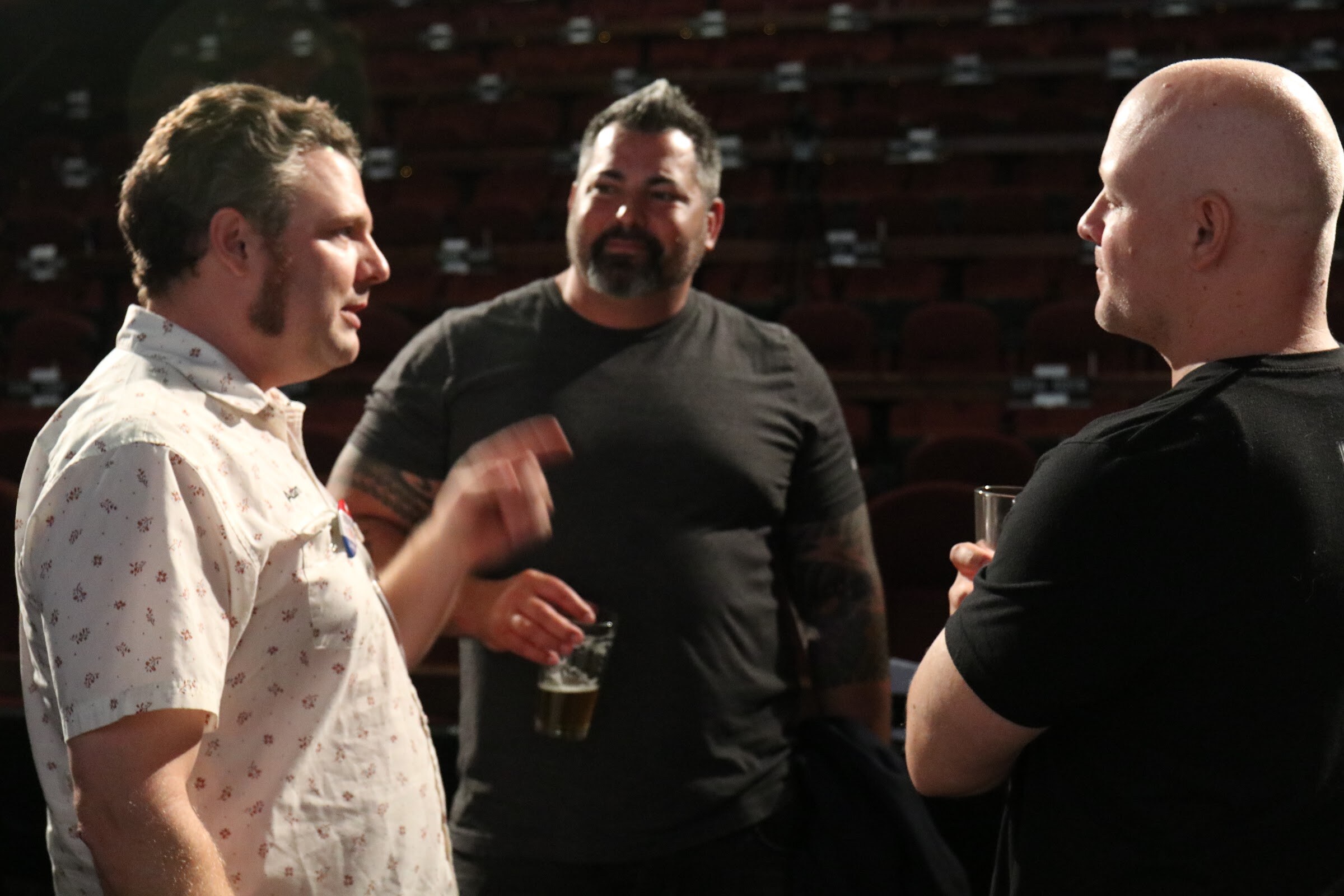 A man in a patterned white shirt gestures as he talks with a bearded man holding a beer and a bald man in a black shirt, with dark theater seats behind them.