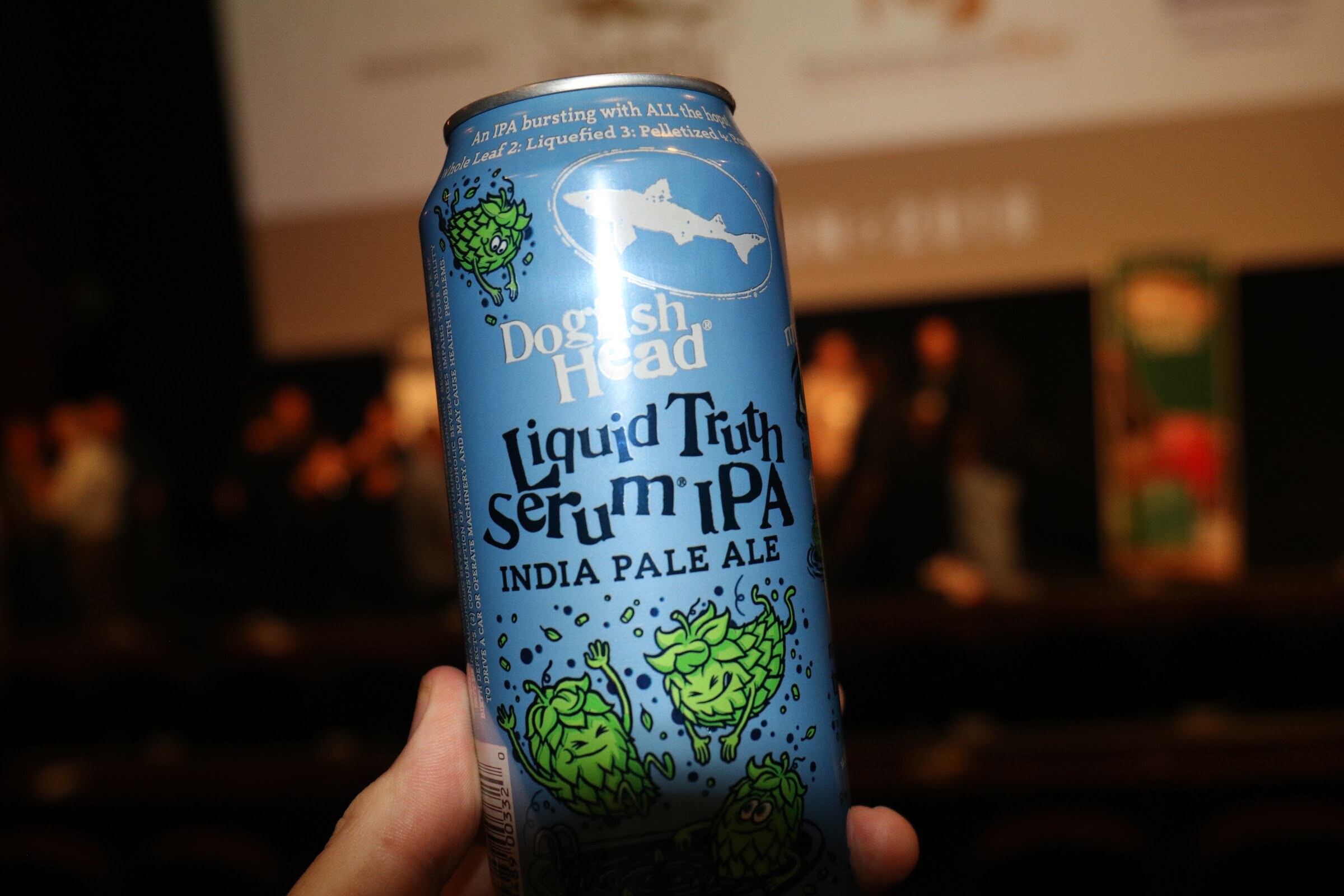 A blue can of Dogfish Head Liquid Truth Serum IPA with cartoon hops held up in front of a blurry cinema stage.