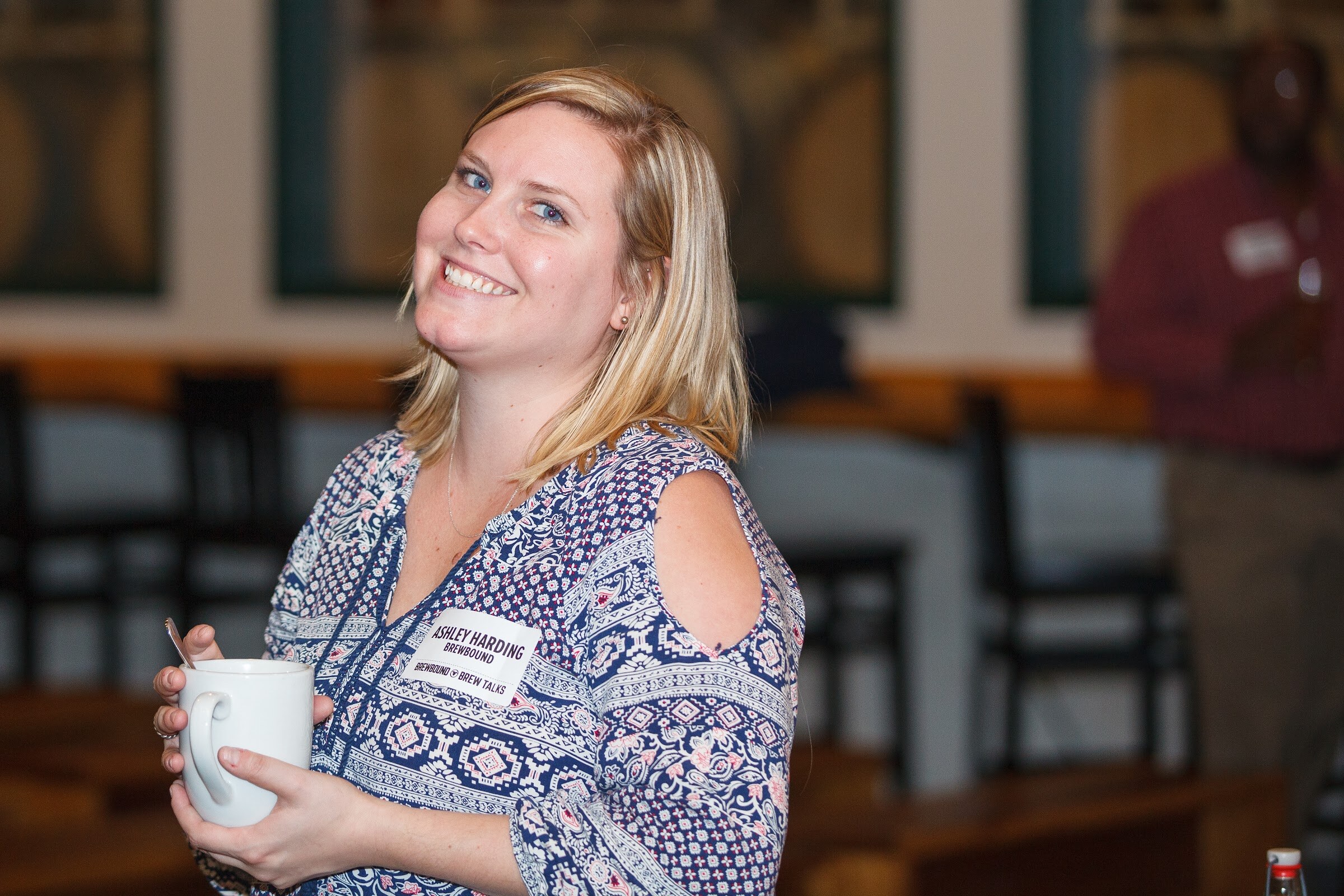 A woman in a blue patterned blouse smiles at the camera while holding a white mug.