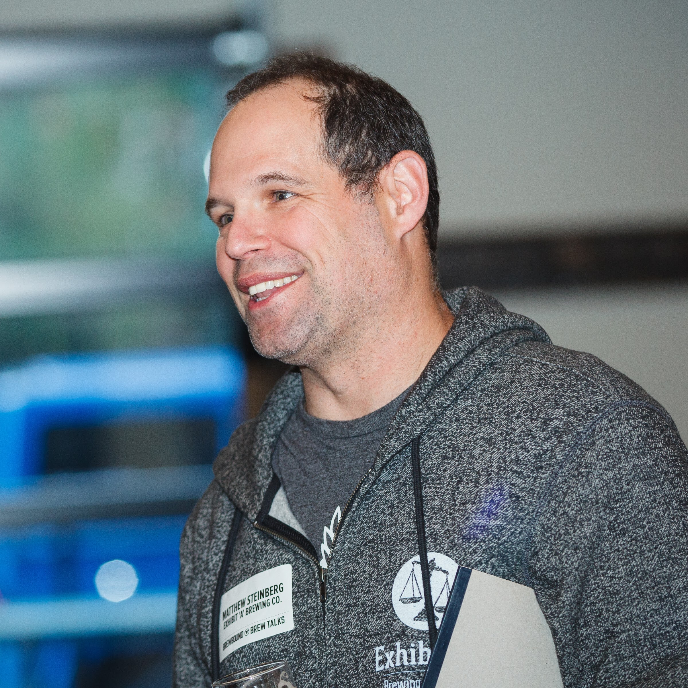 A man in a gray zippered hoodie wears an event badge and smiles to the side.