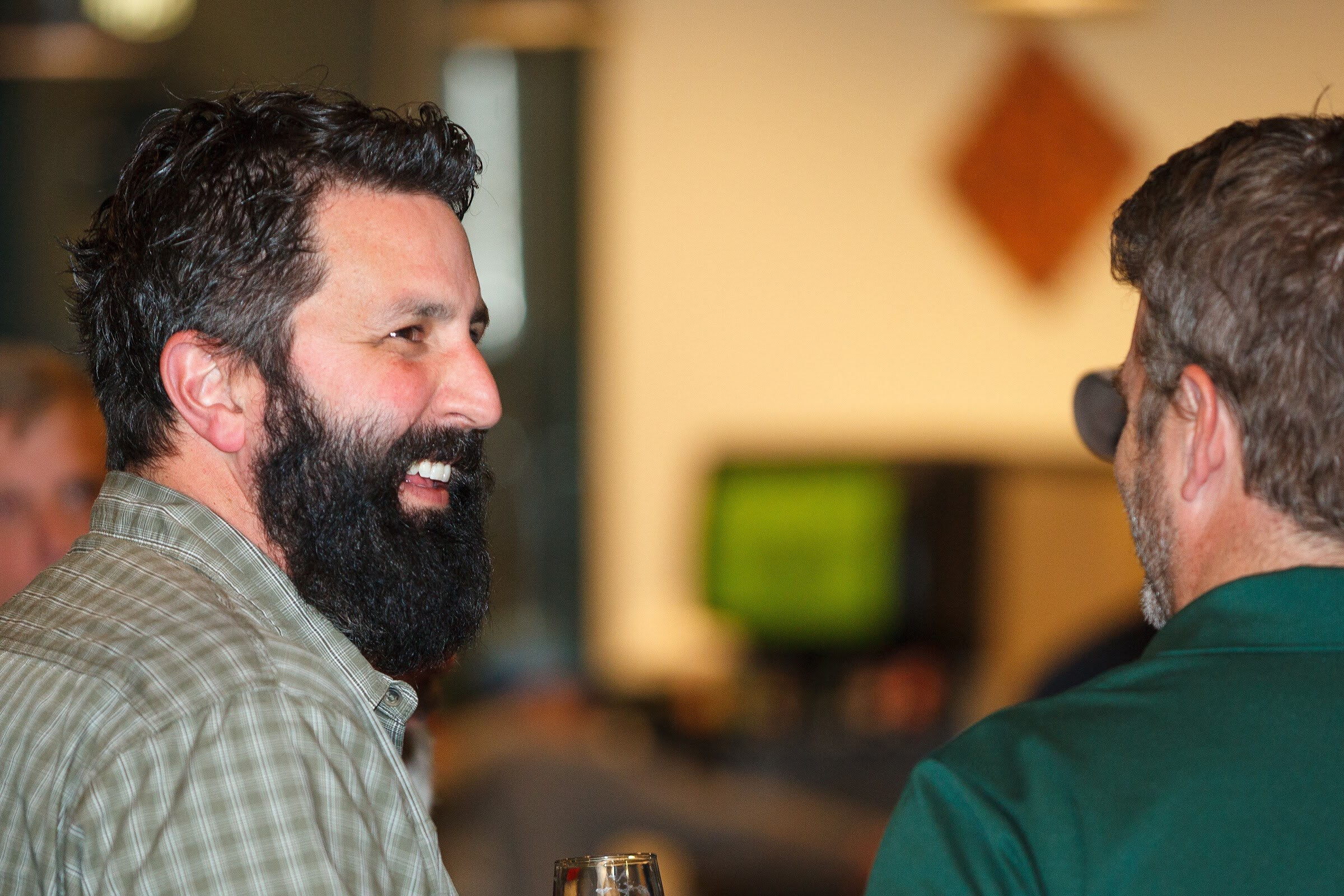 A bearded man in a plaid shirt smiles at a man in a green shirt, with a beer glass between them.