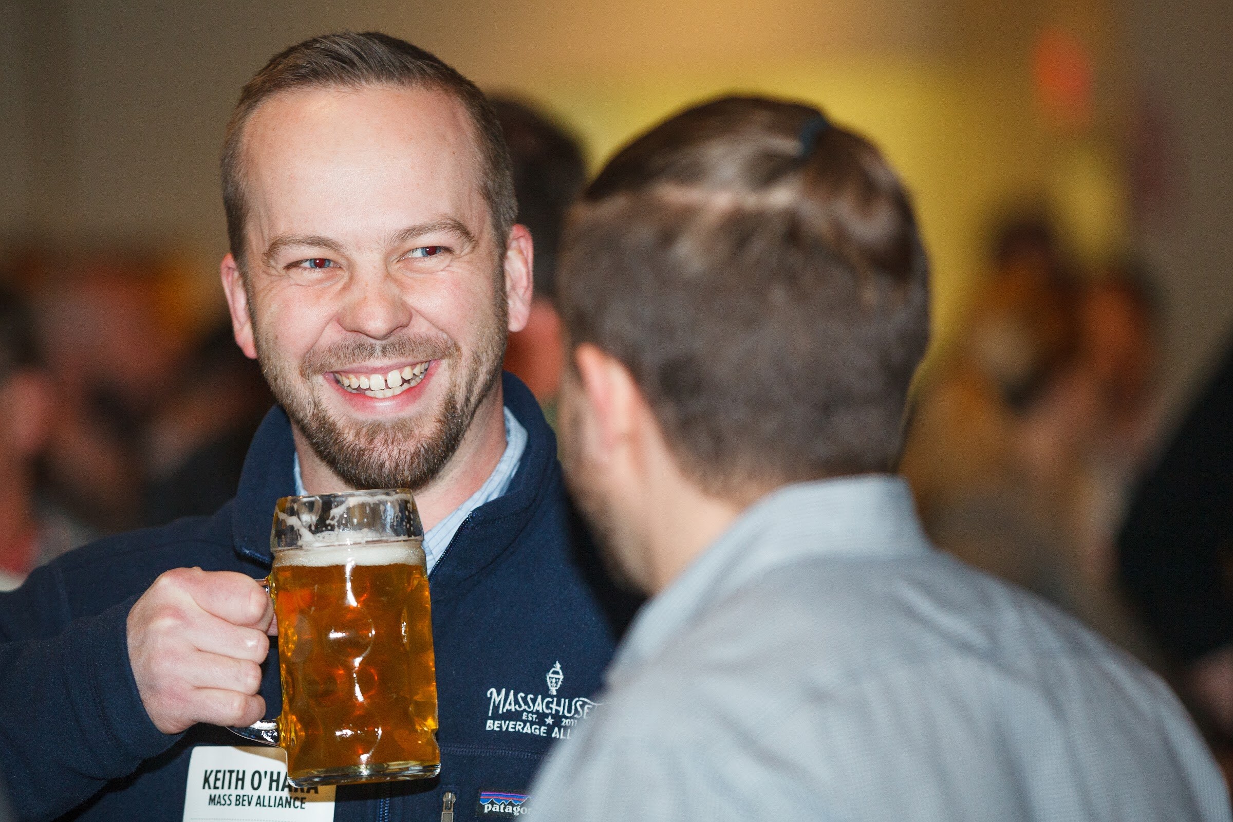 A smiling man holds a large glass mug of amber beer while facing another attendee.