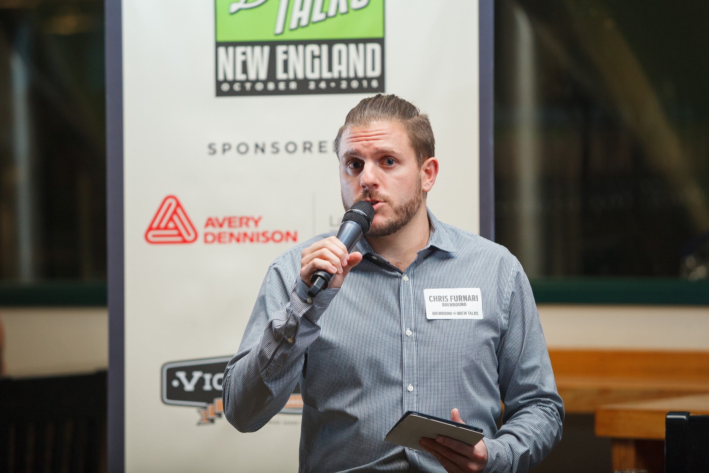 A man holds a microphone and a tablet beside a Brew Talks New England sign bearing sponsor logos.