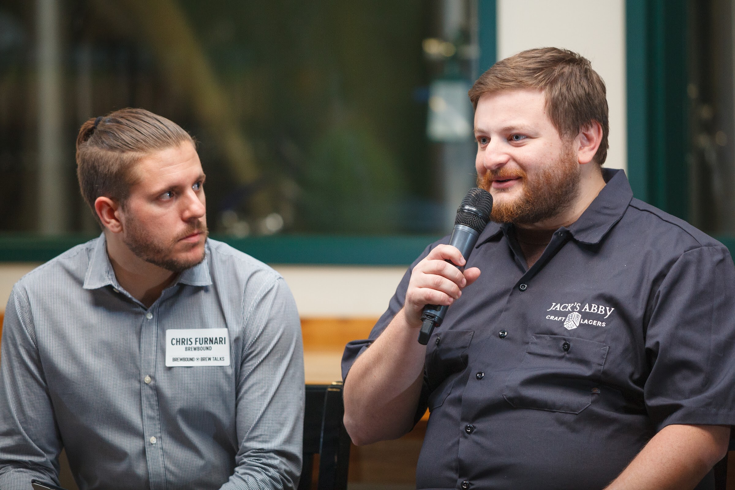 Two men sit in front of brewery windows, with the man in a dark Jack’s Abby shirt holding a microphone.