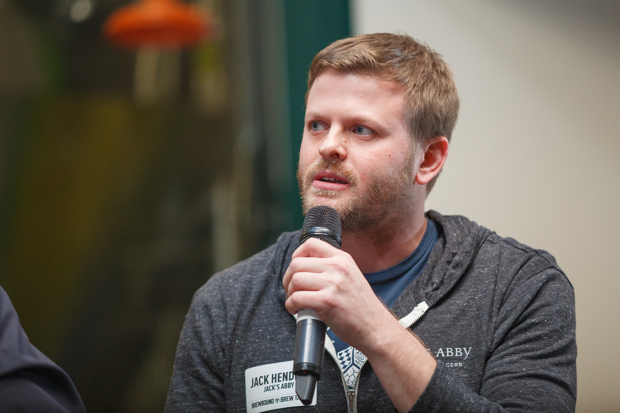 A man wearing a gray Jack’s Abby hoodie holds a microphone near his mouth.