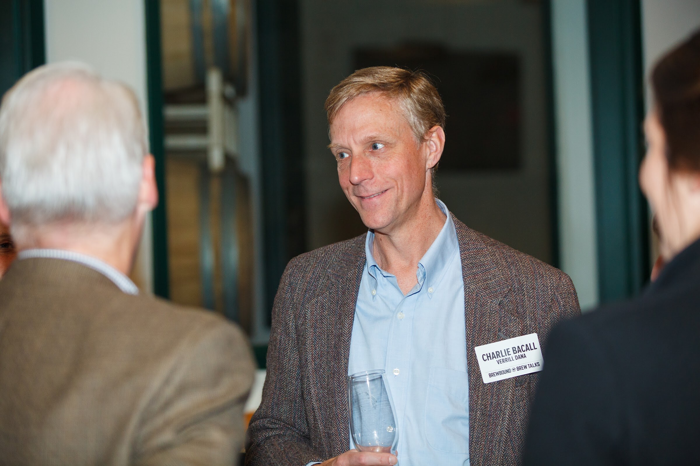 A man in a brown jacket holds an empty glass while speaking with two people at the edges of the frame.