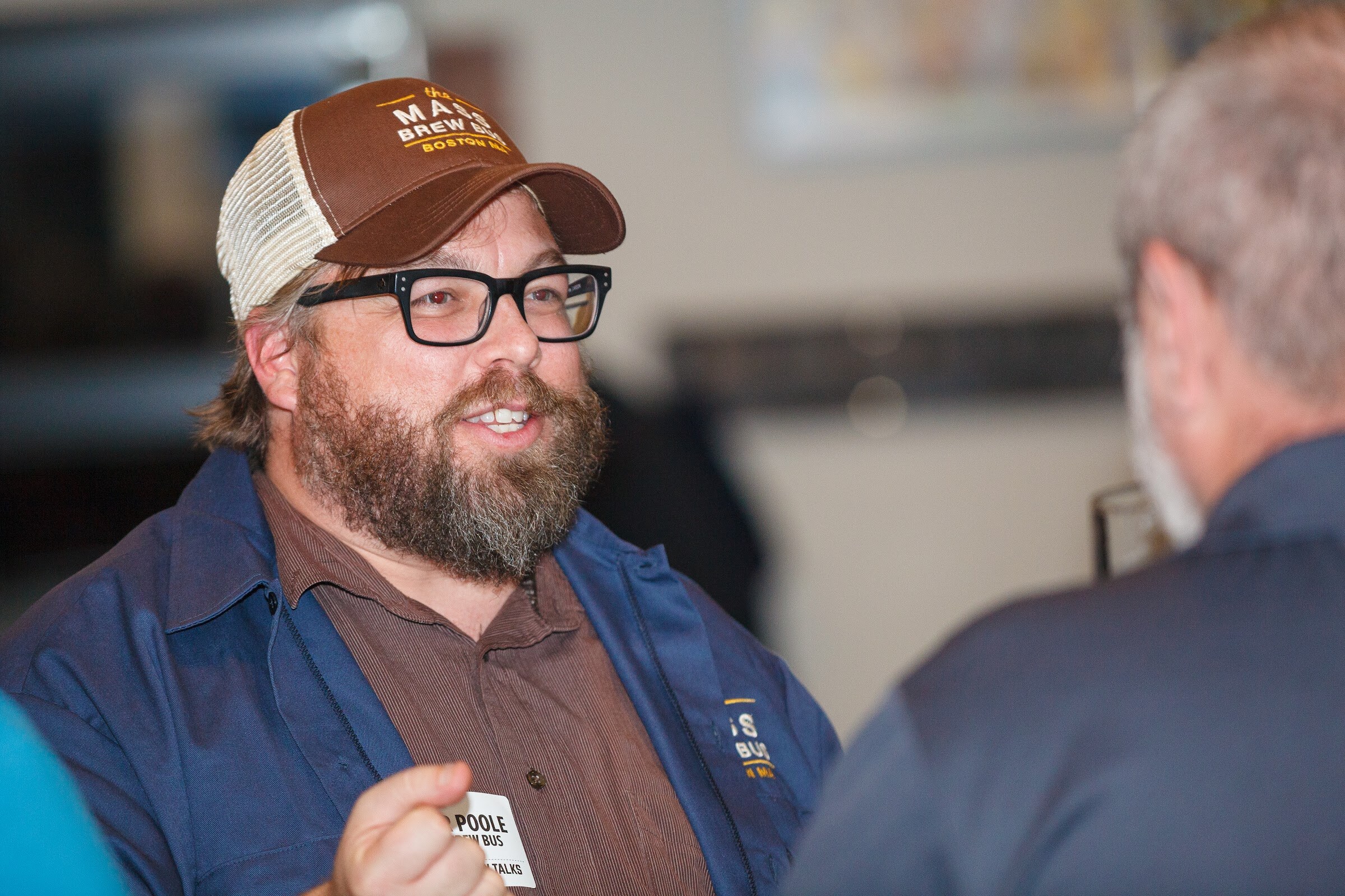 A bearded man wearing glasses and a brown cap raises a hand during a conversation.