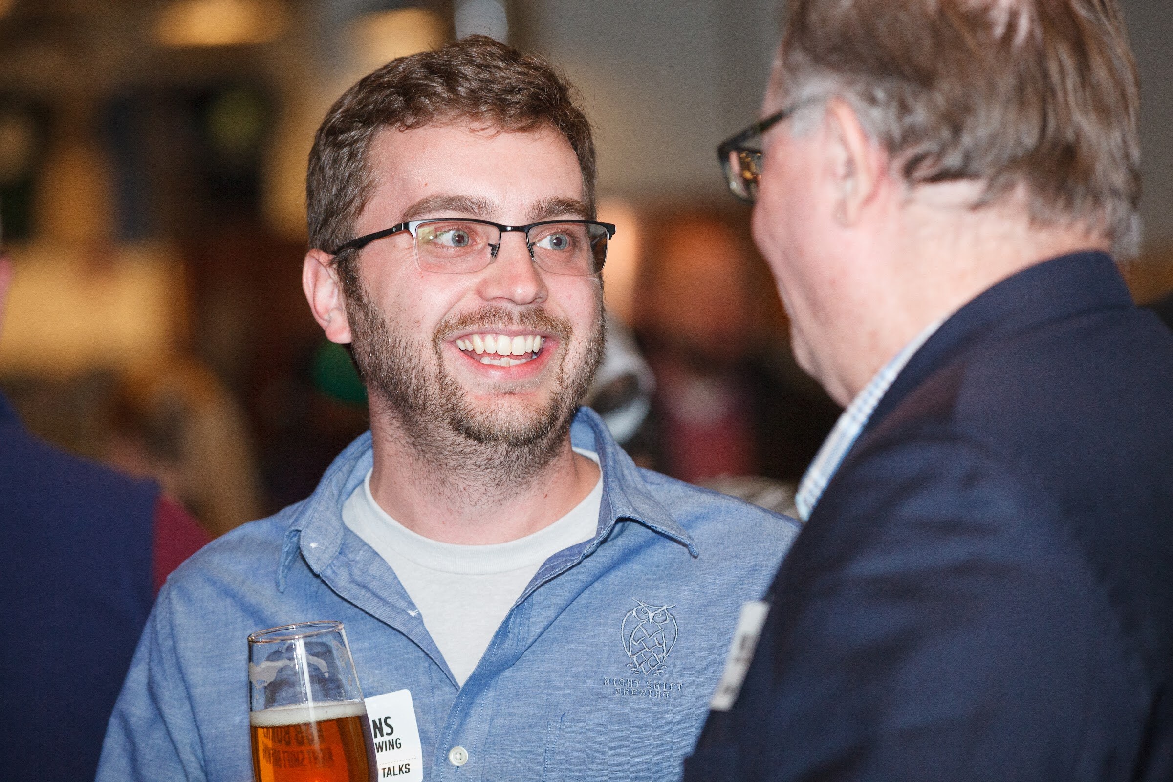 A man in glasses and a blue shirt holds a tall beer glass while facing a man in a dark jacket.