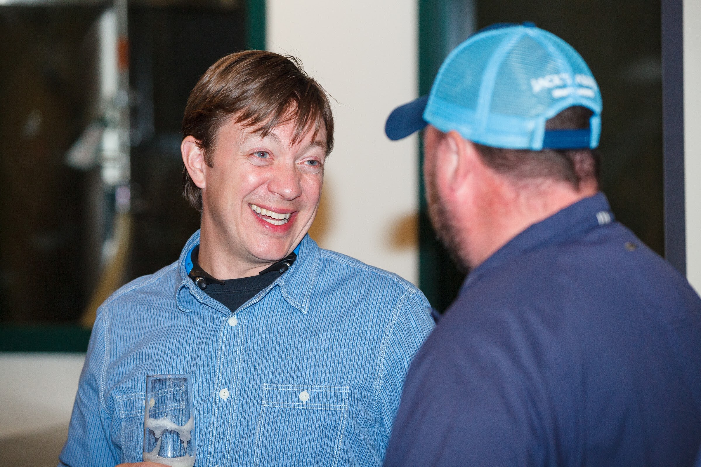 A man in a light blue shirt holds a beer glass and smiles at a man wearing a blue cap.