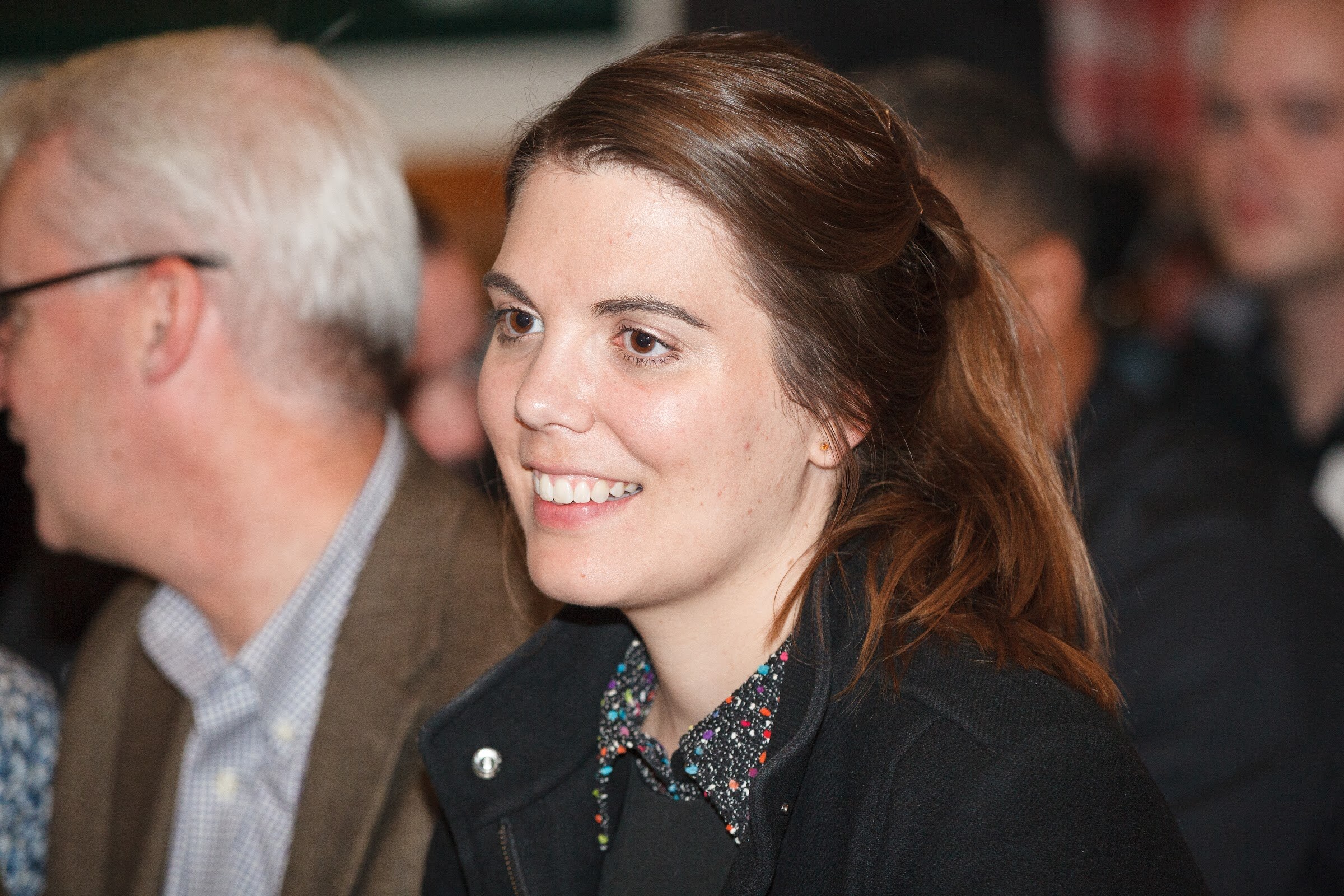 A woman in a black jacket smiles in the audience beside a man wearing glasses.