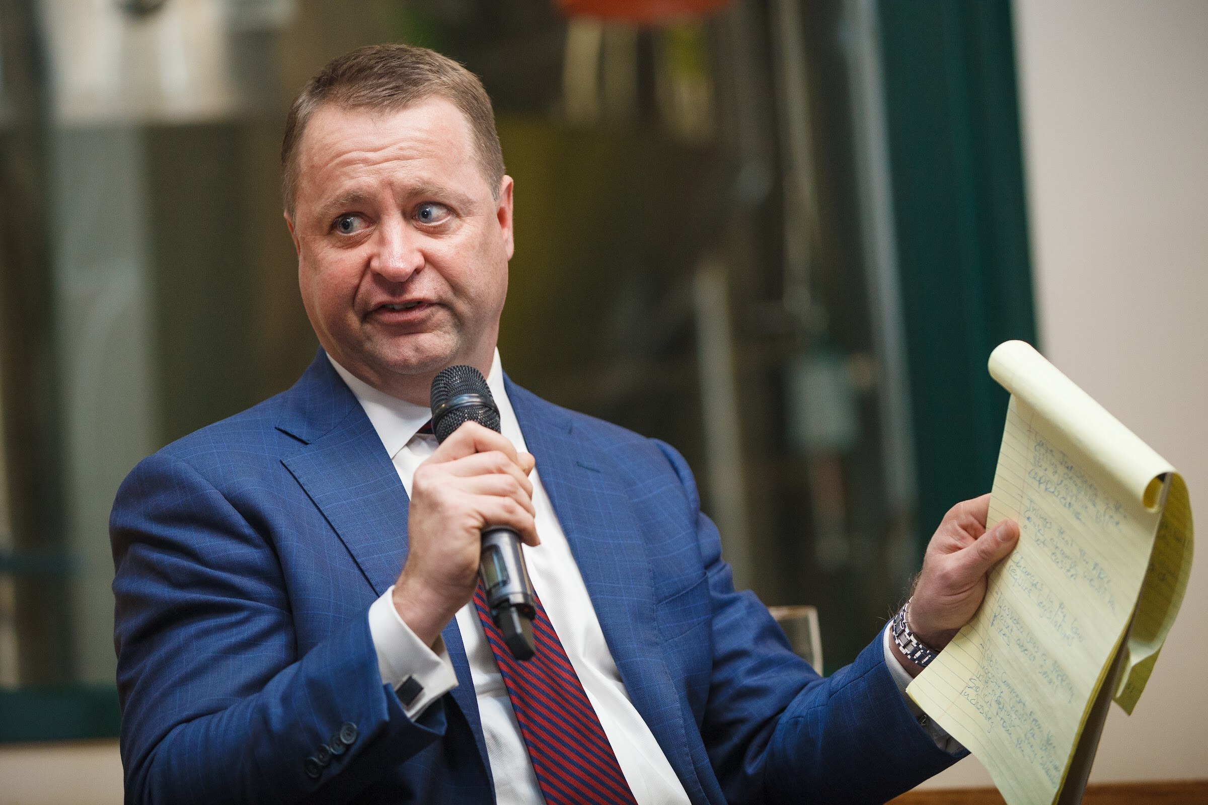 A man in a blue suit holds a microphone in one hand and a yellow notepad in the other.