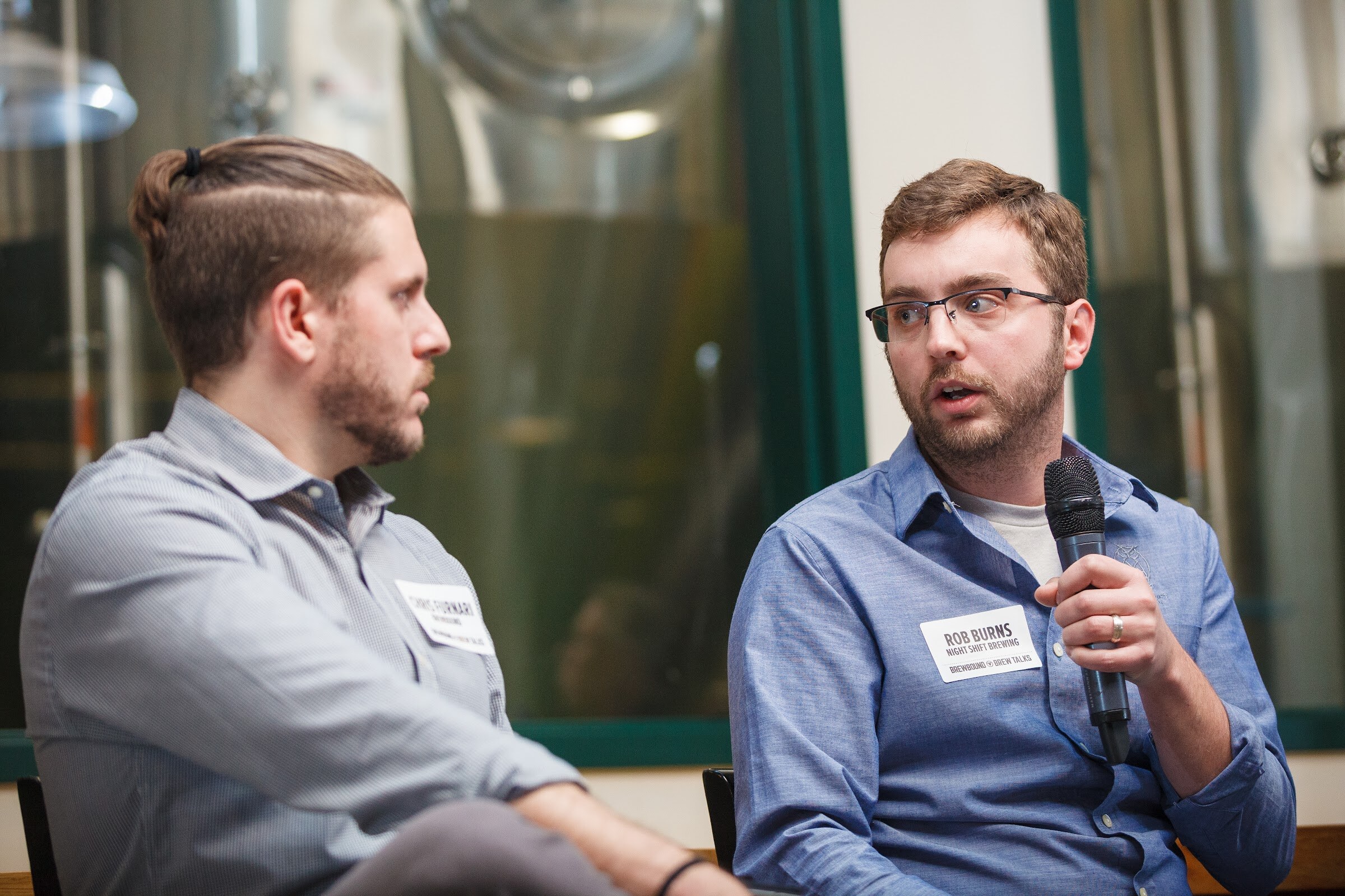 A man in glasses and a blue shirt holds a microphone while seated beside a man in a gray shirt.