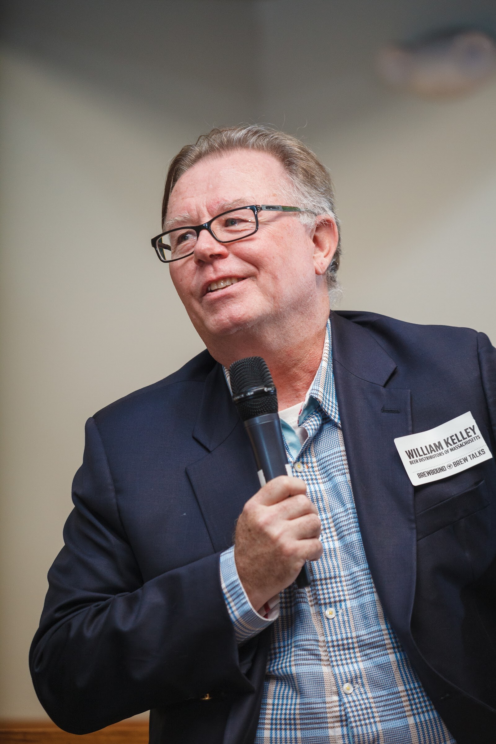 A man in glasses, a plaid shirt and a dark jacket sits holding a microphone.
